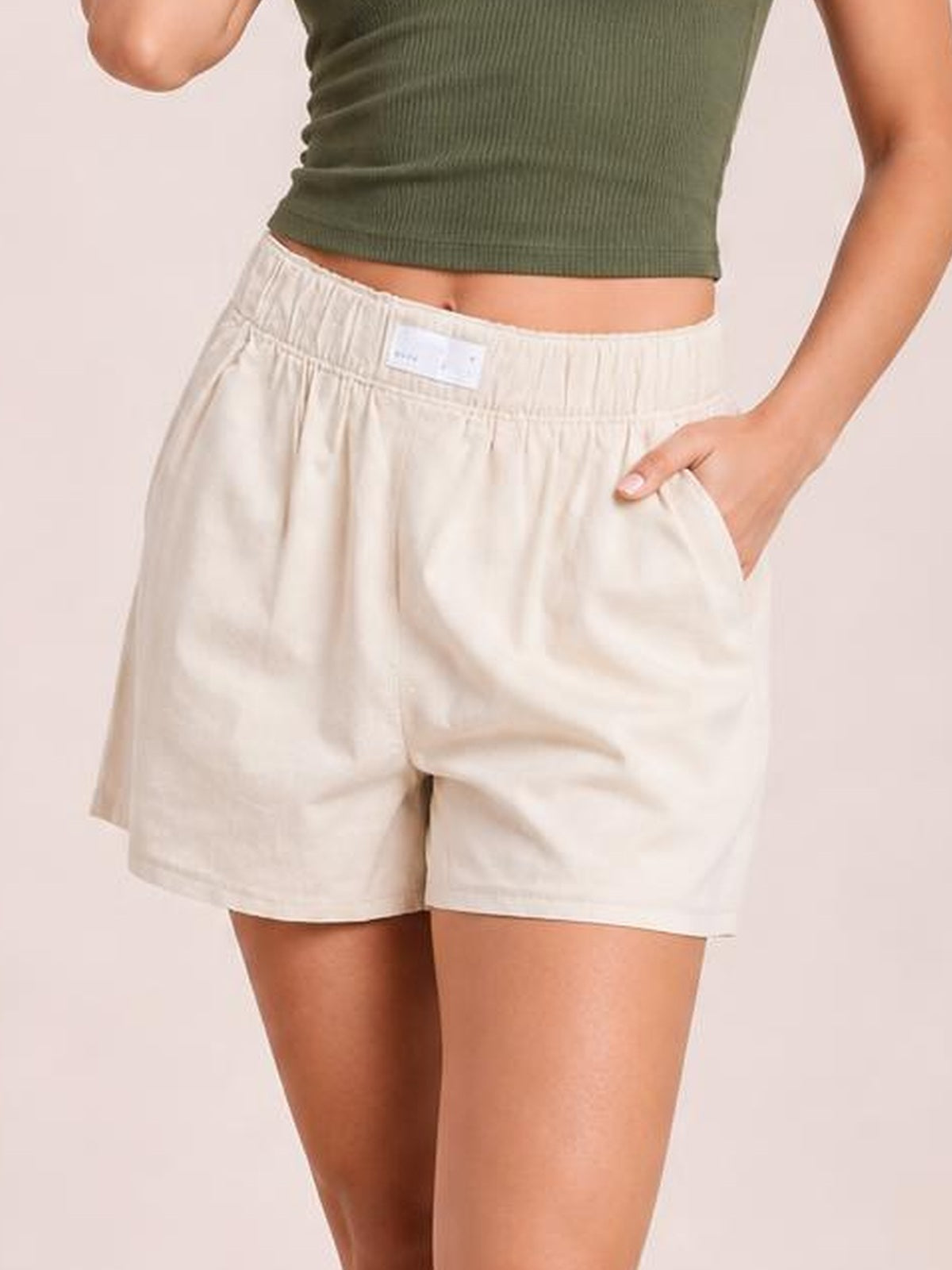 Model wearing relaxed cream elastic waist shorts with a mid-rise waistband, roomy silhouette, and side pockets, posed with one hand tucked into a pocket.
