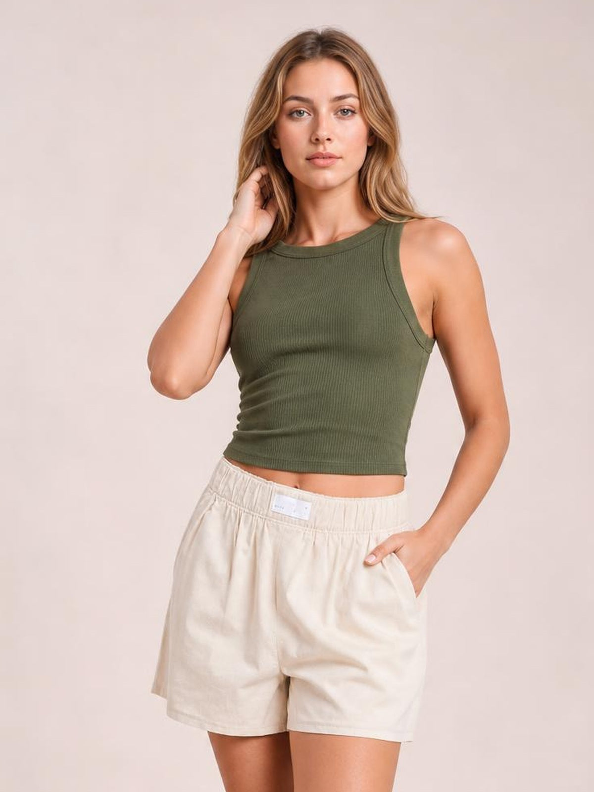 A woman modeling a ribbed olive green cropped tank top and cream-colored linen shorts against a solid light beige studio background.