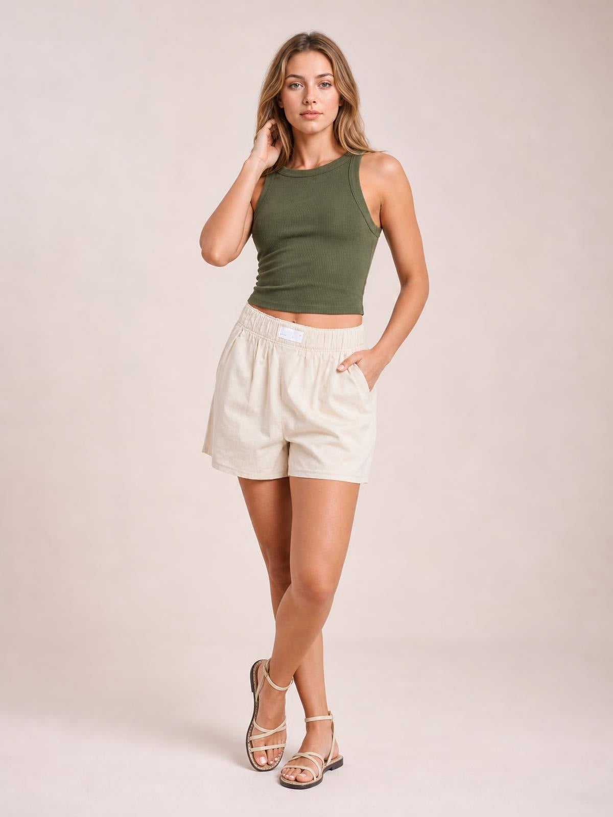 A model poses with one hand in the pocket, wearing cream elastic waist shorts with a relaxed mid-rise silhouette, side pockets, and straight hems.