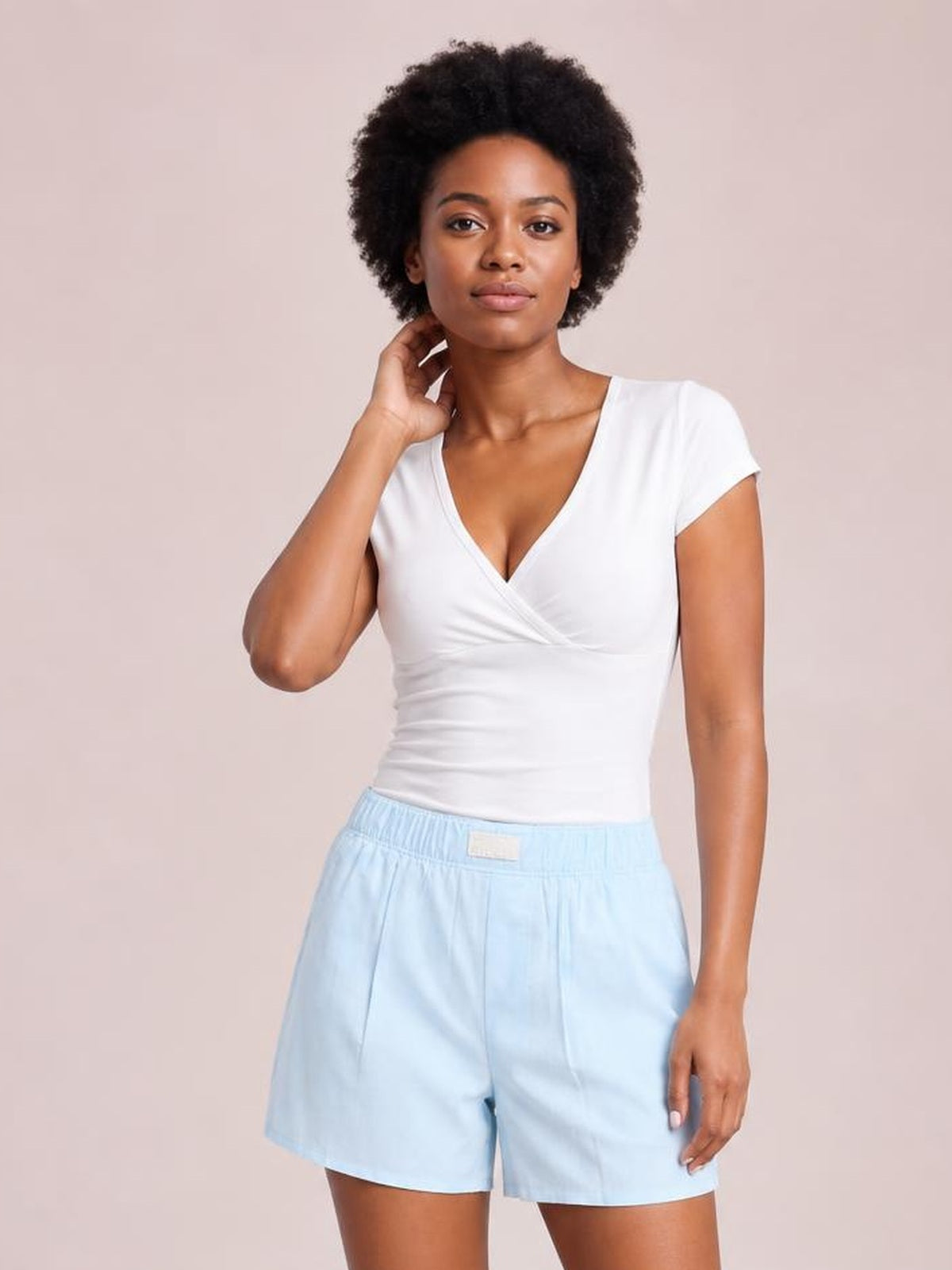 Model standing with one hand near her neck, wearing light blue elastic waist shorts with a relaxed straight-leg silhouette, front pockets, and a white fitted top.