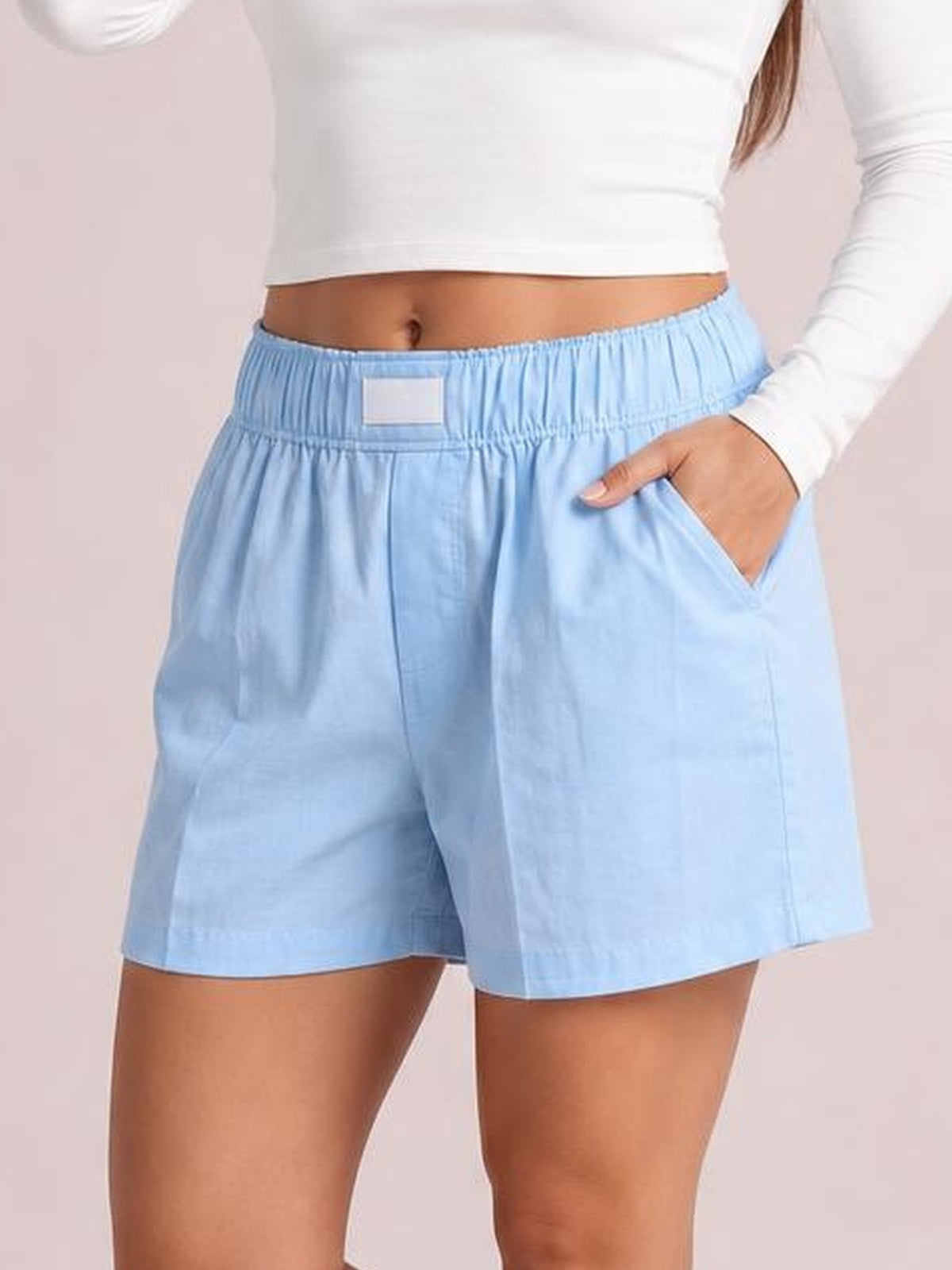 A model wears light blue elastic waist shorts with a relaxed, loose silhouette, straight hems, and a convenient side pocket shown with one hand inside.
