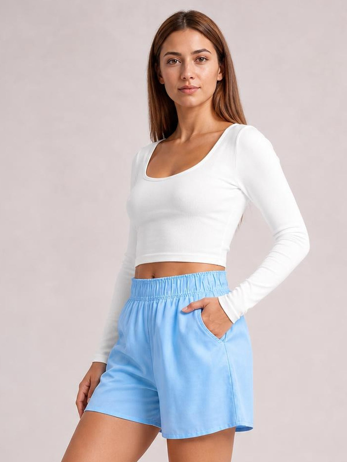 A woman modeling a white long-sleeve crop top and light blue shorts against a plain light beige studio background.