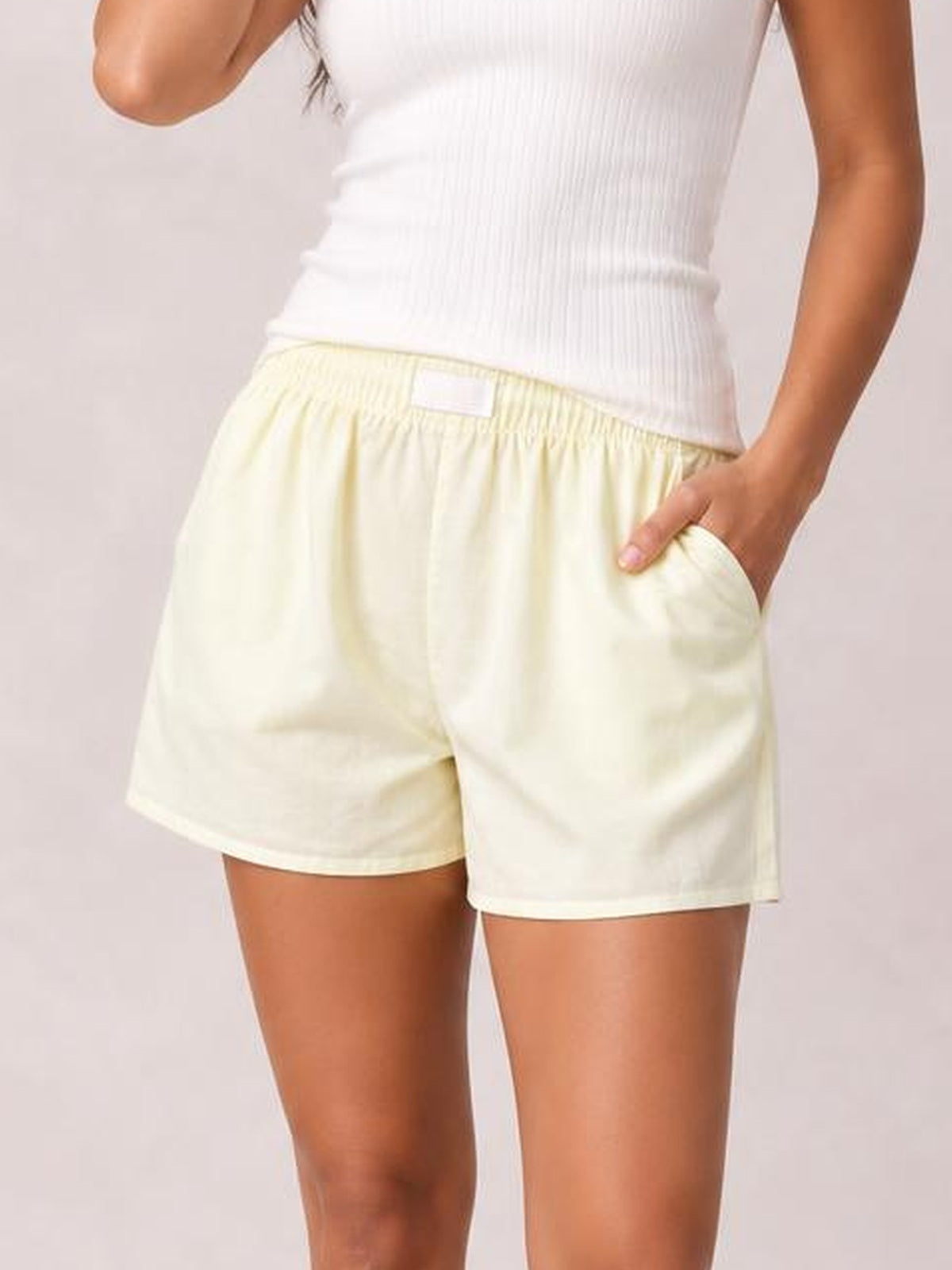 Model wears pale yellow elastic waist shorts with a relaxed silhouette, straight hems, and convenient side pockets, shown with one hand tucked into a pocket.