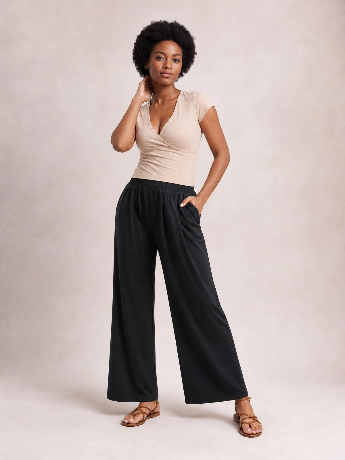 Model poses with one hand in a pocket, wearing black elastic waist wide leg pants with a fluid full-length silhouette and subtle front pleats.
