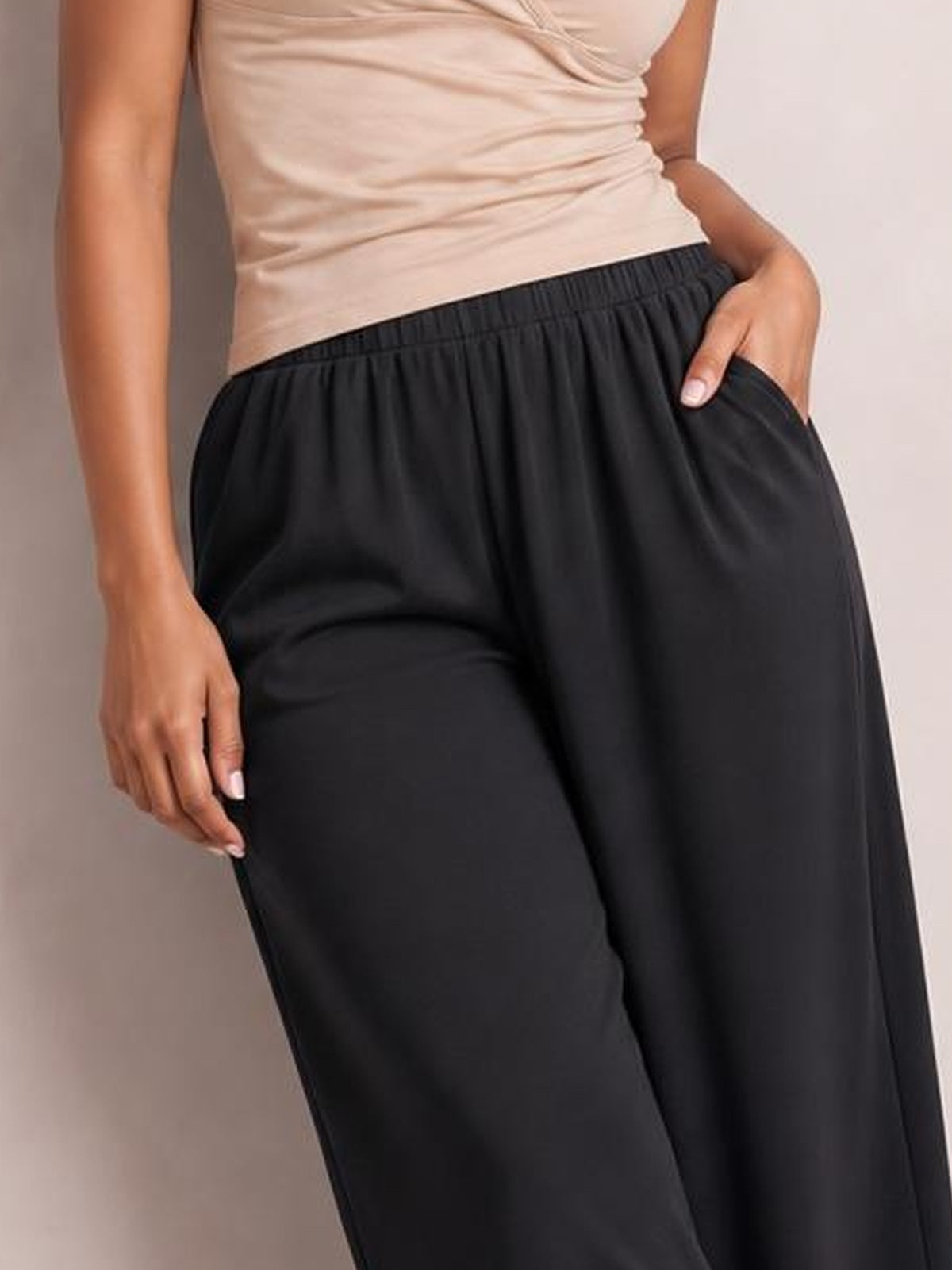 A model wears black elastic-waist wide-leg pants with a relaxed silhouette and side pockets, posing with one hand tucked into a pocket.