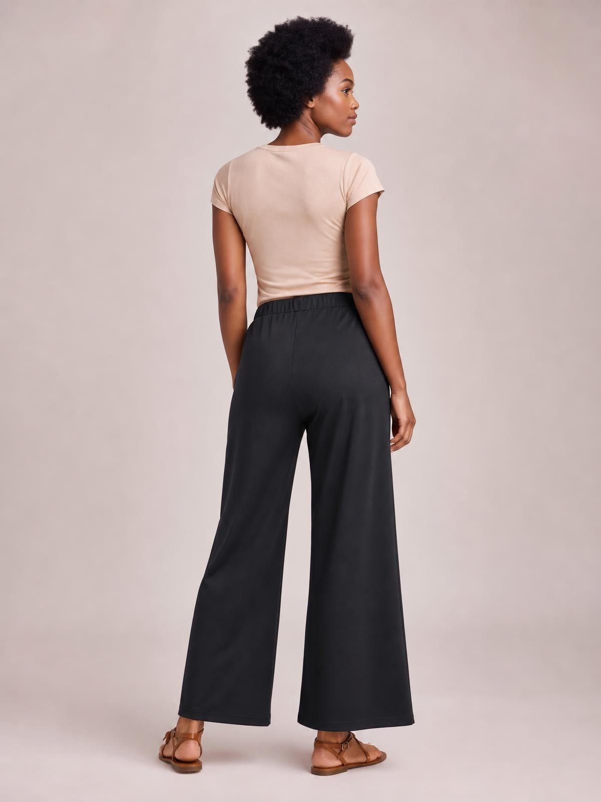 A model poses from behind in black elastic-waist wide-leg pants with side pockets, a smooth opaque finish, and a relaxed full-length silhouette.