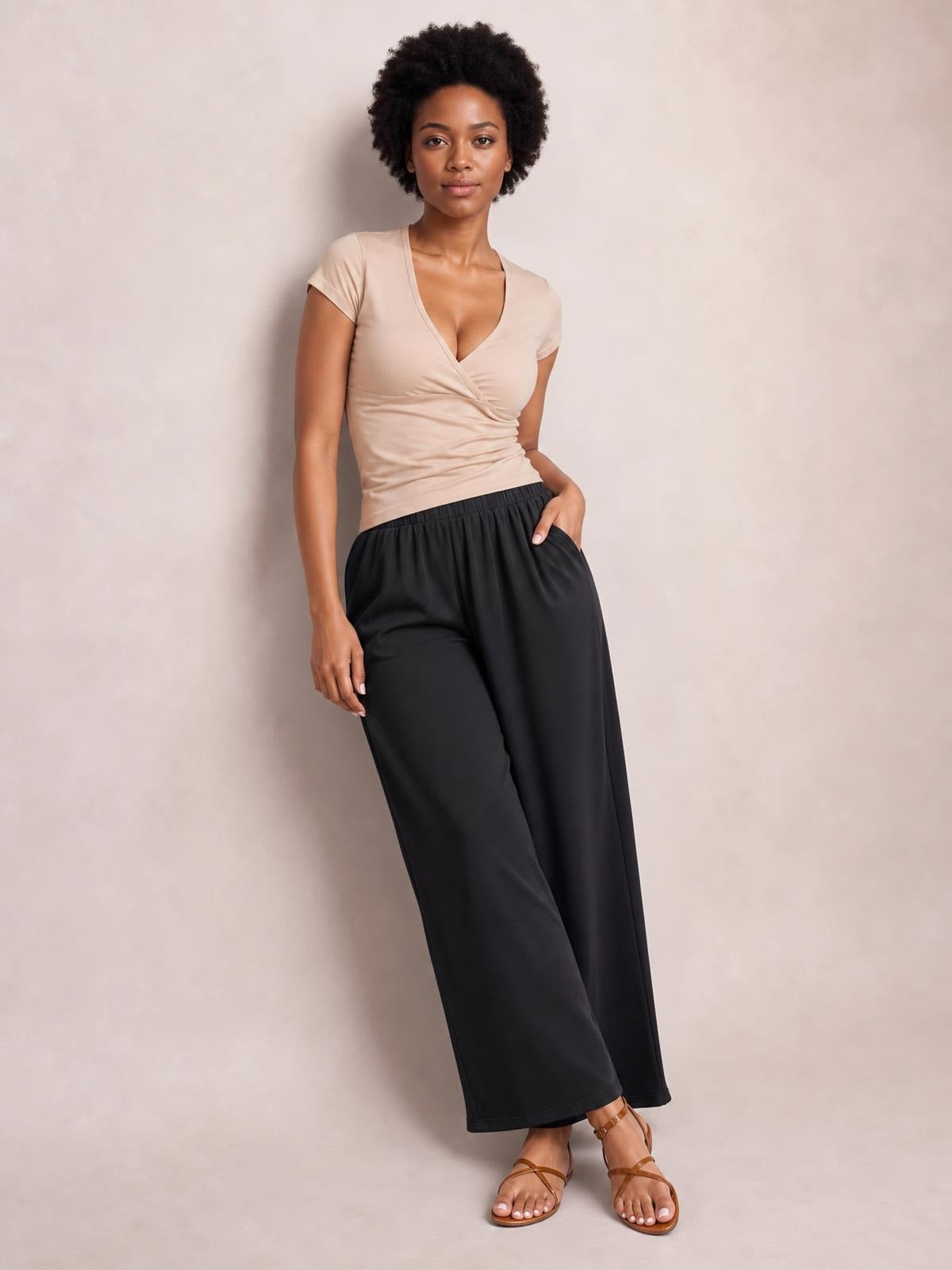 Model wearing black elastic waist wide leg pants with a flowing full-length silhouette and side pockets, posed with one hand in a pocket.