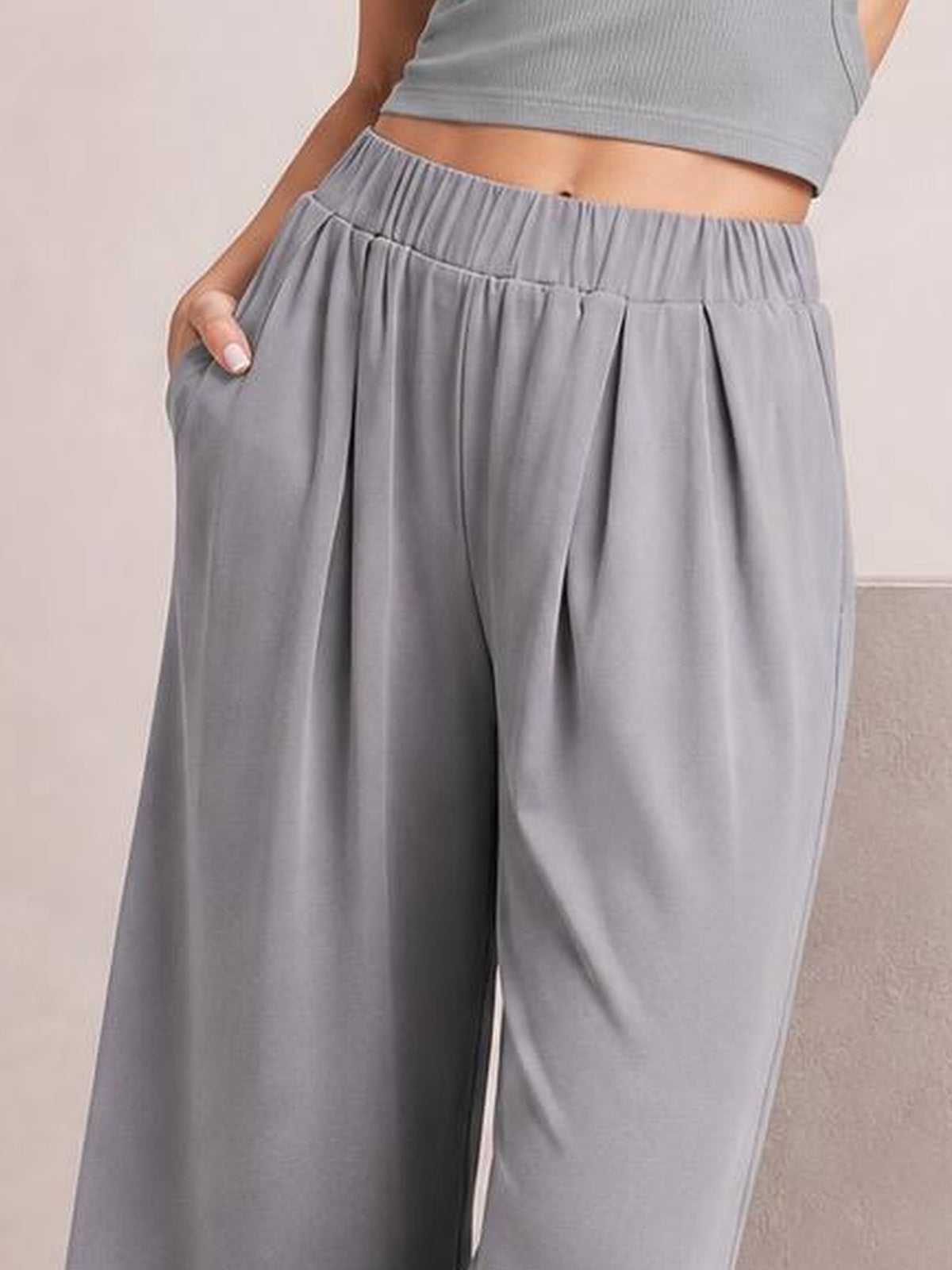 Model wearing gray elastic waist wide leg pants with a flowing silhouette, pleated front construction, and side pockets, shown with one hand tucked into a pocket.