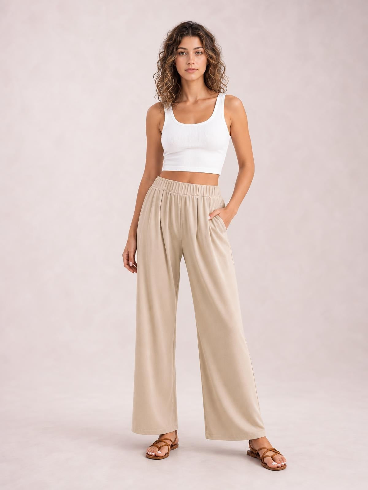 Model poses in beige elastic waist wide leg pants with a flowing full-length silhouette and side pockets, paired with a white cropped tank.