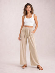 Model poses in beige elastic waist wide leg pants with a flowing full-length silhouette and side pockets, paired with a white cropped tank.