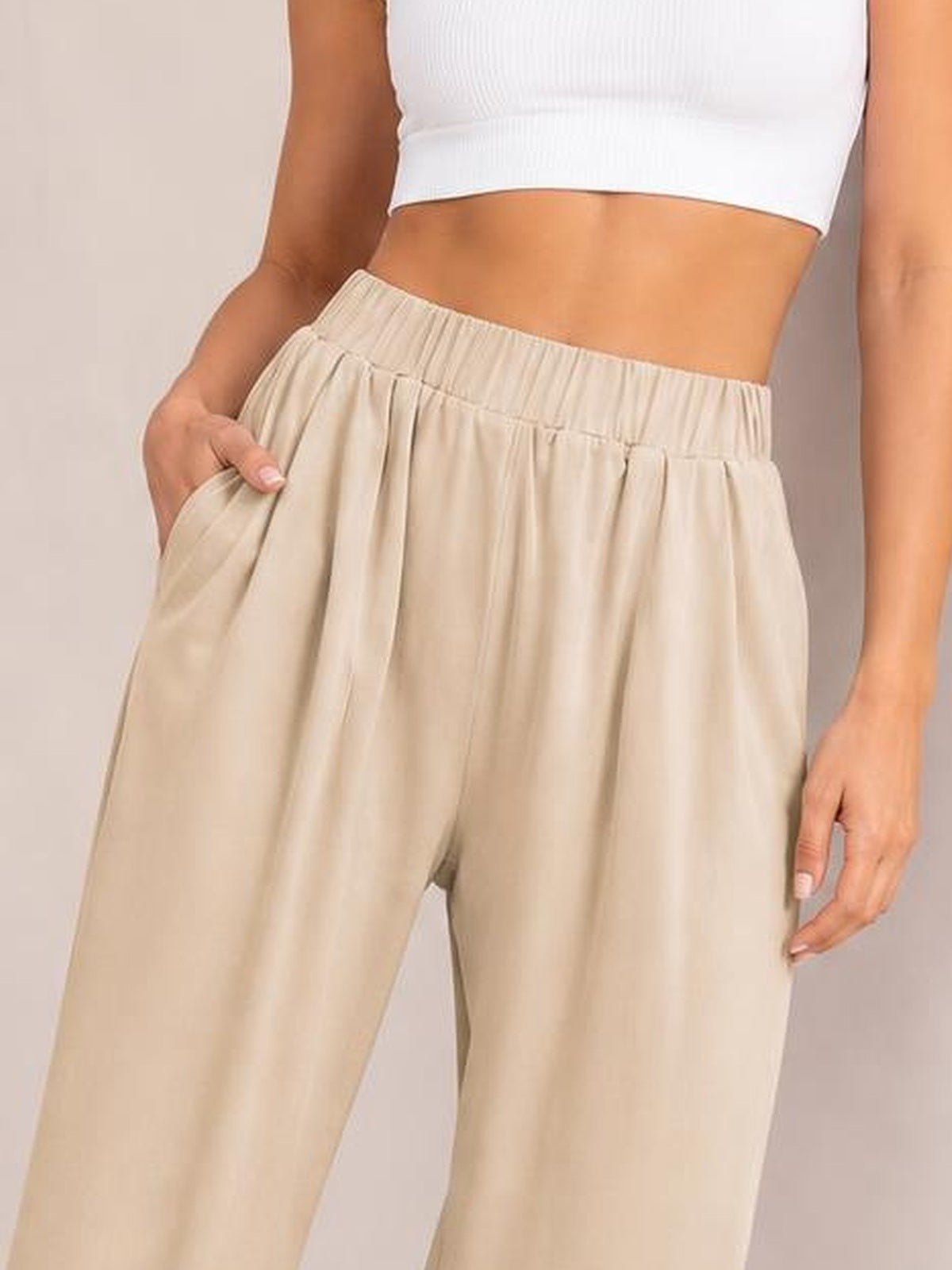 A model wears beige elastic-waist wide-leg pants with a softly gathered waistband, relaxed silhouette, and side pockets, shown with one hand tucked into a pocket.