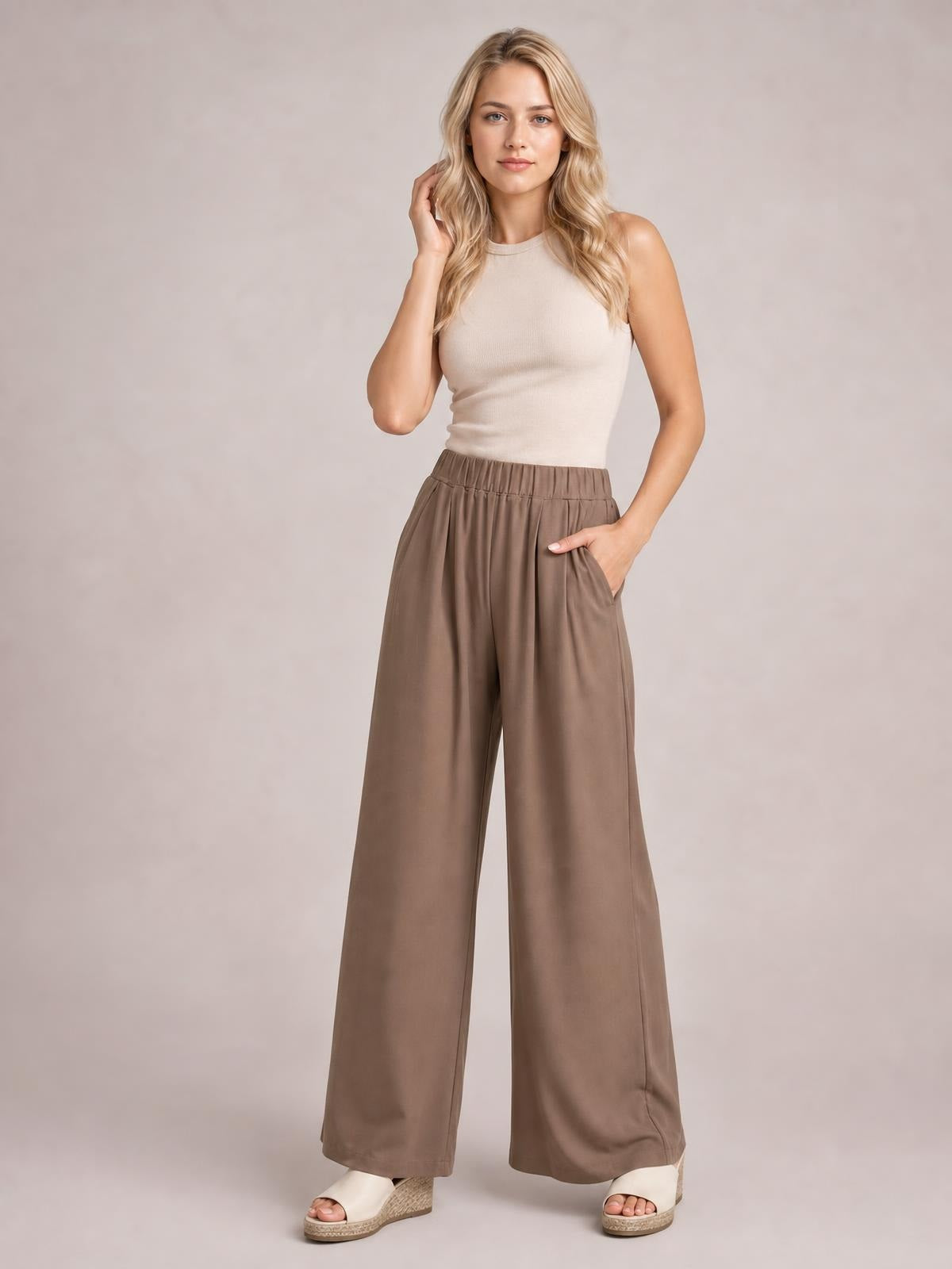 A model poses in taupe elastic waist wide leg pants with a flowing silhouette, visible side pockets, and a hand resting inside one pocket.