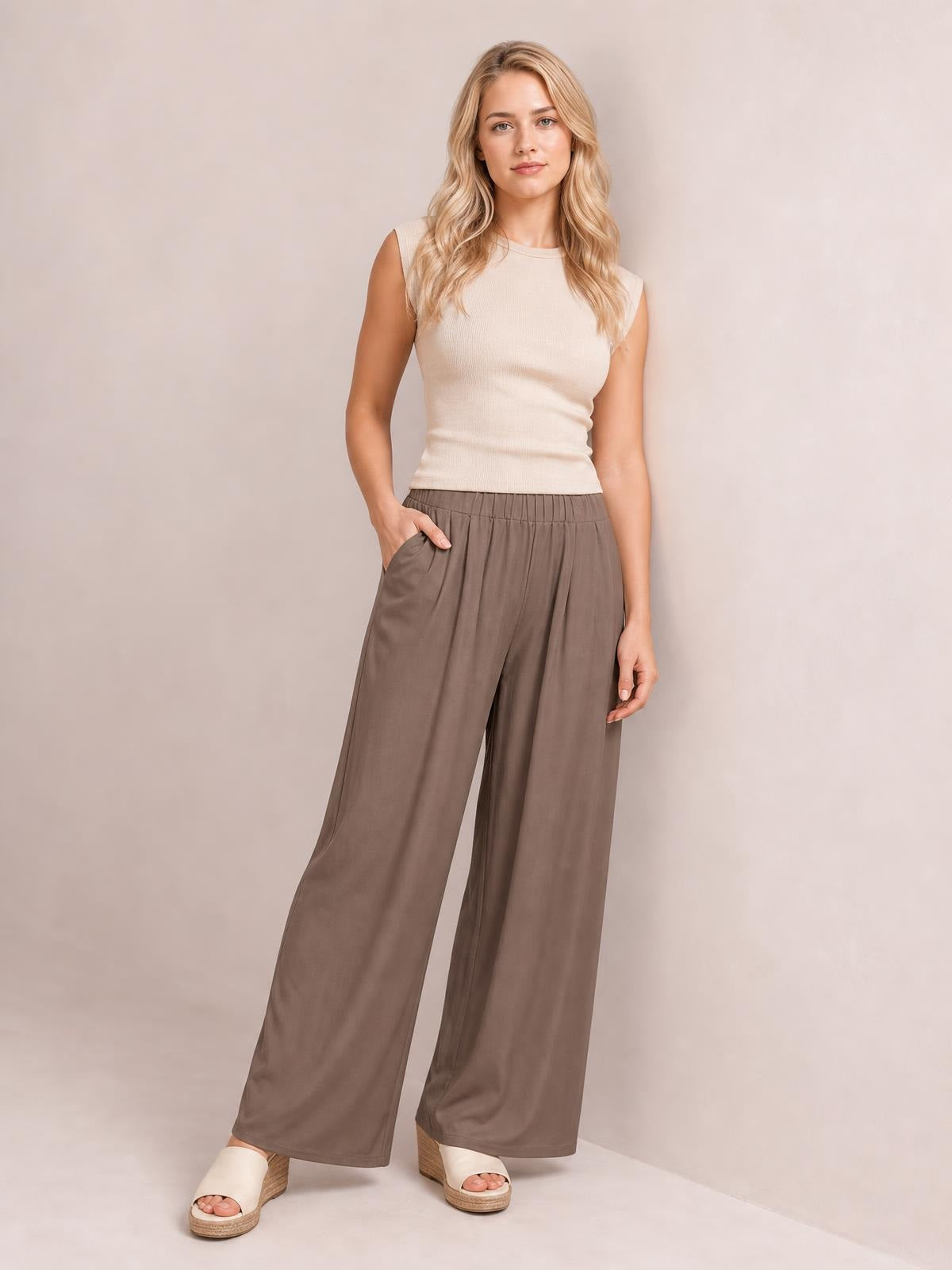 Model stands in taupe elastic waist wide leg pants with a relaxed full-length silhouette, soft drape, and side pockets, one hand tucked into a pocket.