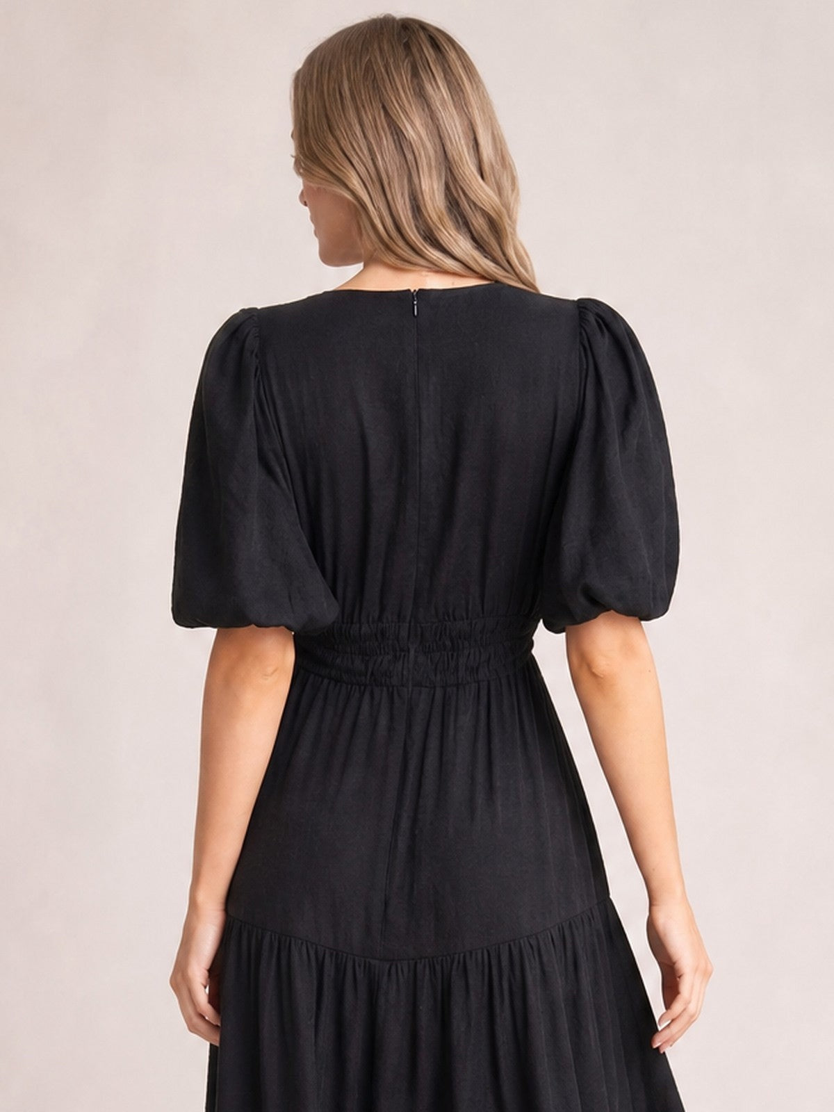 A model stands facing away in a black midi dress with voluminous puff sleeves, a smocked waist, center-back zipper, and flowing tiered skirt.