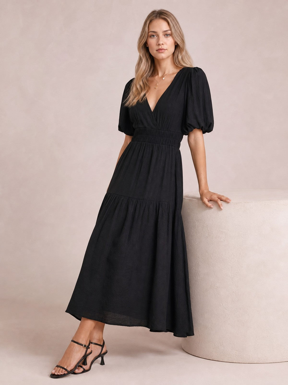 A model leans against a pedestal in an elegant black V-neck midi dress with puff sleeves, a smocked waist, and a flowing tiered skirt.