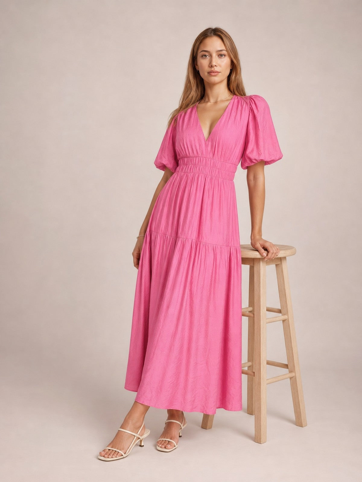 Model poses beside a stool in a bright pink puff sleeve V-neck midi dress with a smocked waist, tiered skirt, and flowing A-line silhouette.