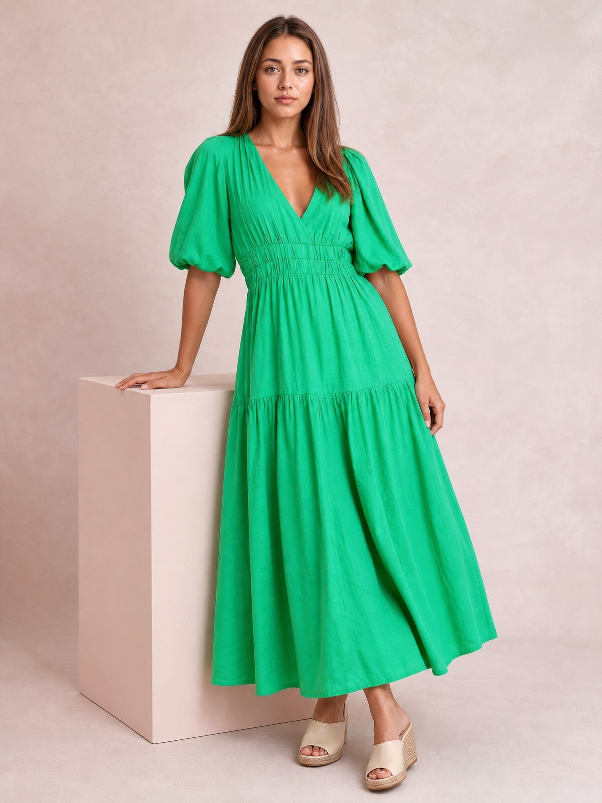 Model poses beside a pedestal in a bright green V-neck midi dress with puff sleeves, a smocked waist, and a flowing tiered skirt.