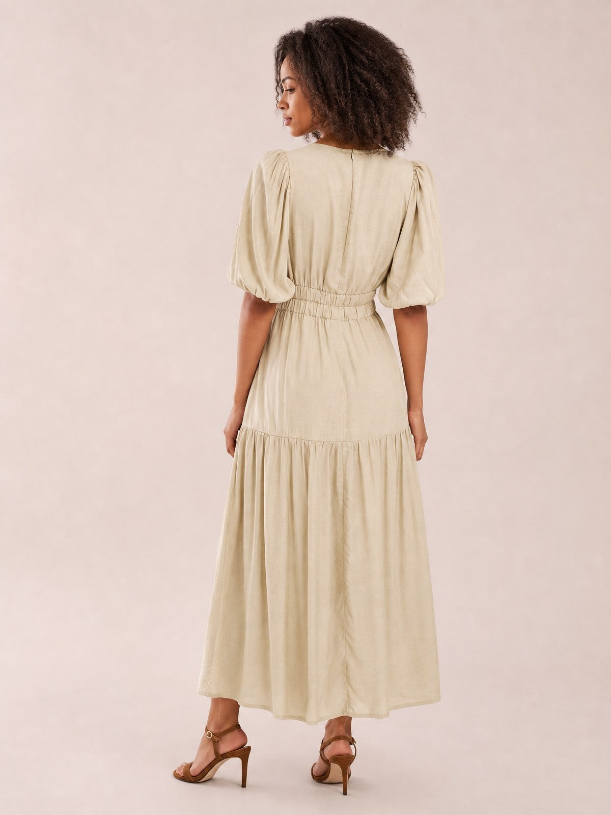 Model poses with her back turned in an elegant beige midi dress featuring puff sleeves, a smocked waist, and a flowing tiered skirt.