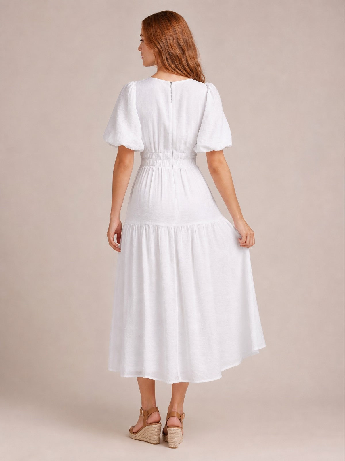 A model poses from behind in a white midi dress with gathered puff sleeves, a smocked waist, and a flowing tiered skirt held gently at the sides.