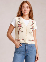 Model wears a cream embroidered button-up sweater vest with a V-neck, floral stitching, front buttons, and ribbed trim over a white T-shirt, posing with one hand in her jeans pocket.