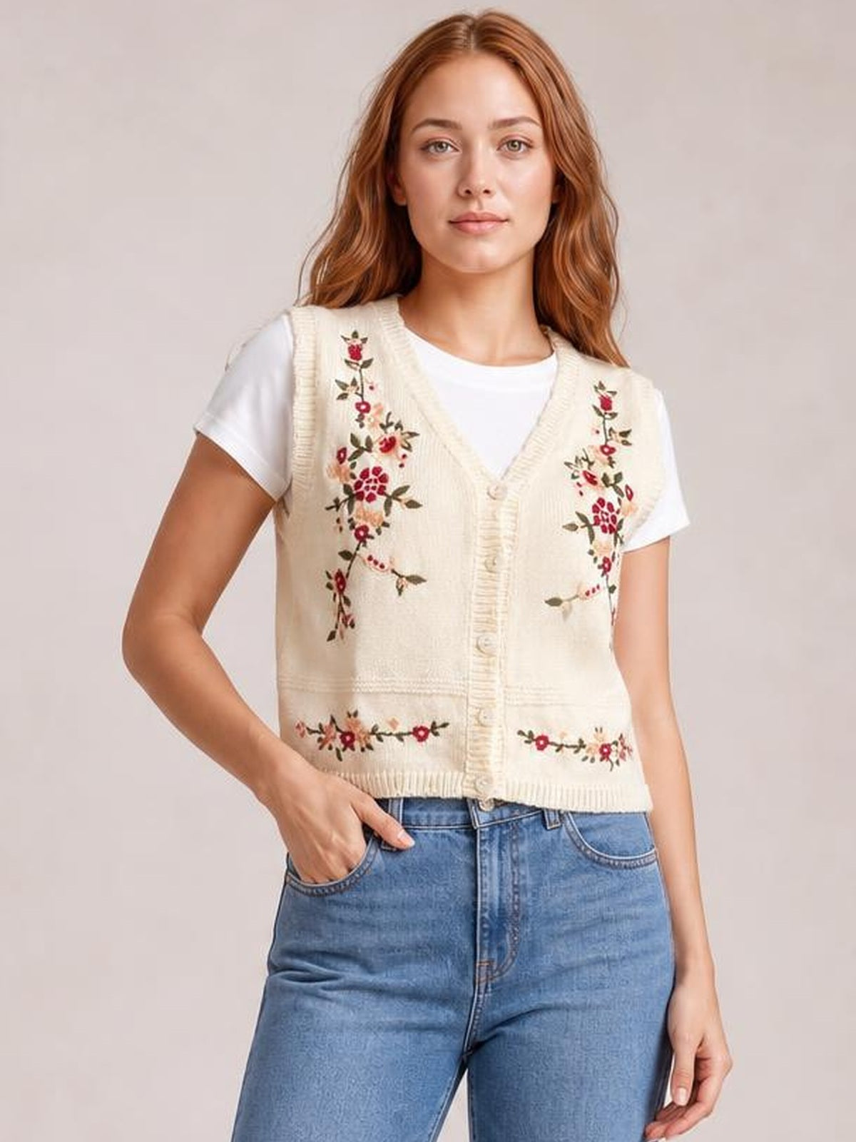 Model wears a cream embroidered button-up sweater vest with a V-neck, floral stitching, front buttons, and ribbed trim over a white T-shirt, posing with one hand in her jeans pocket.