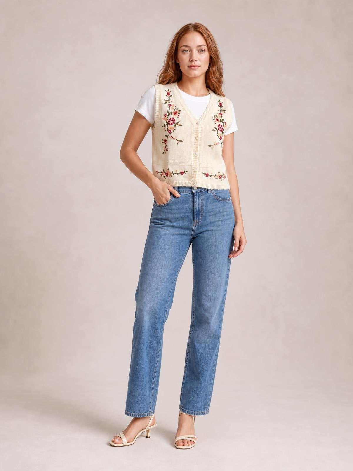 A woman stands in a cream embroidered button-up sweater vest with a V-neck, floral detailing, ribbed trim, and a fitted sleeveless silhouette over a white T-shirt.