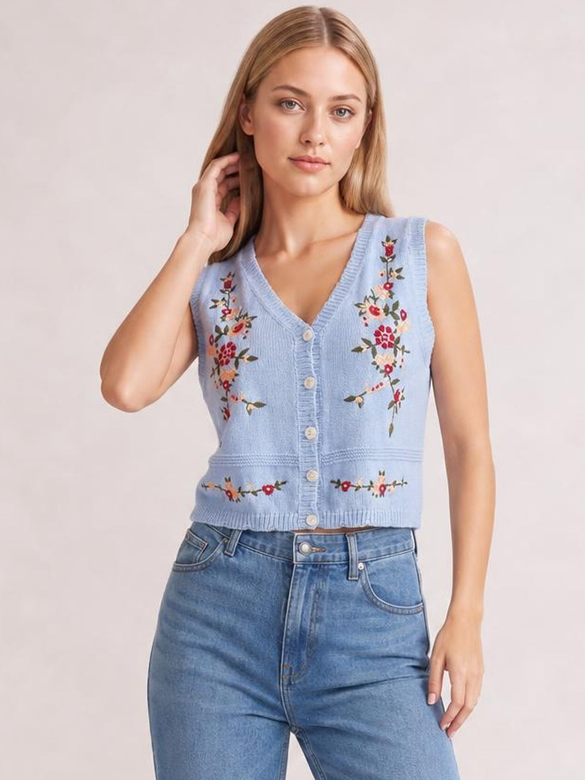 Model poses with one hand in her hair while wearing a light blue embroidered button-up sweater vest with a V-neck, fitted silhouette, and ribbed trim.