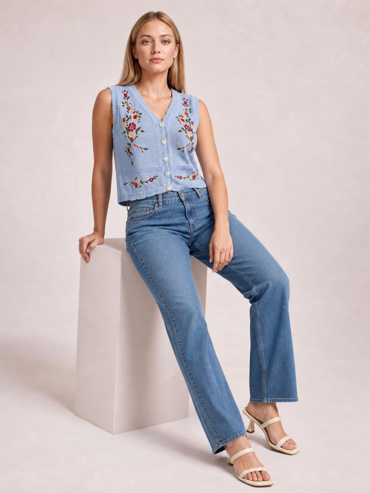 Model seated in a light blue embroidered button-up sweater vest with a V-neck, floral detailing, front buttons, and a cropped straight silhouette.