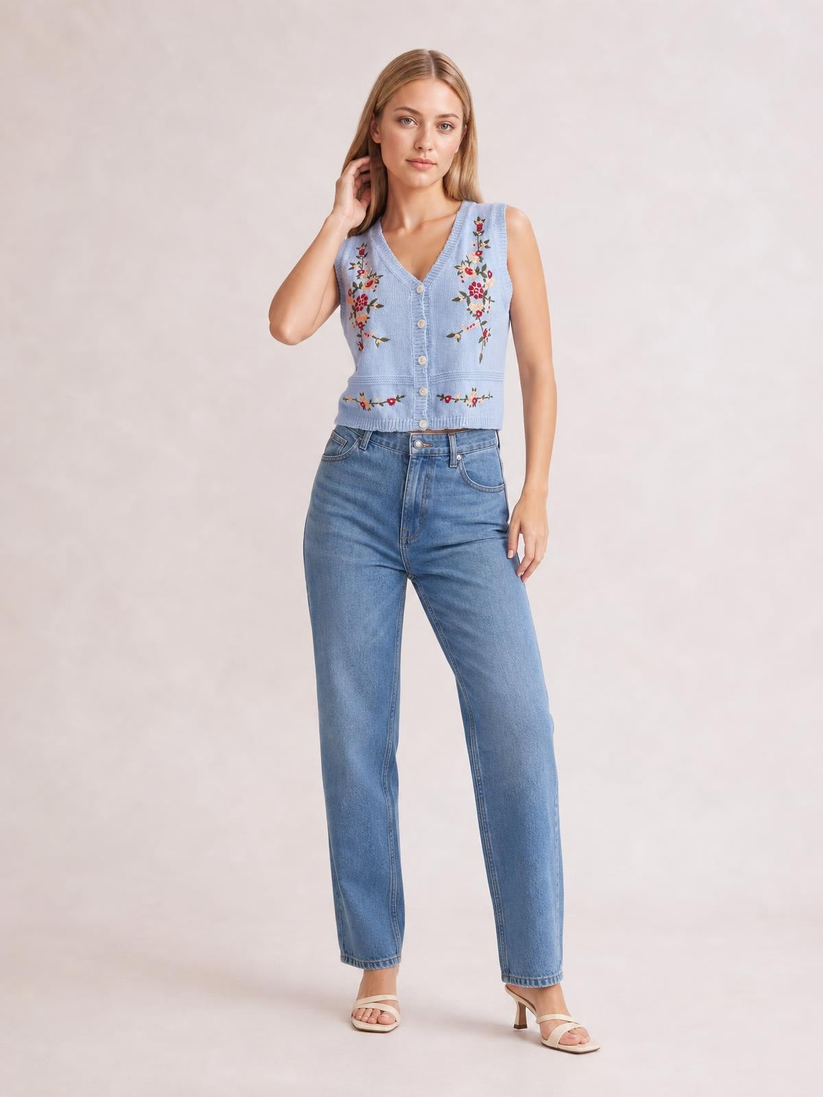 Model poses in a light blue embroidered button-up sweater vest with a V-neck, front buttons, floral detailing, and a fitted cropped silhouette.