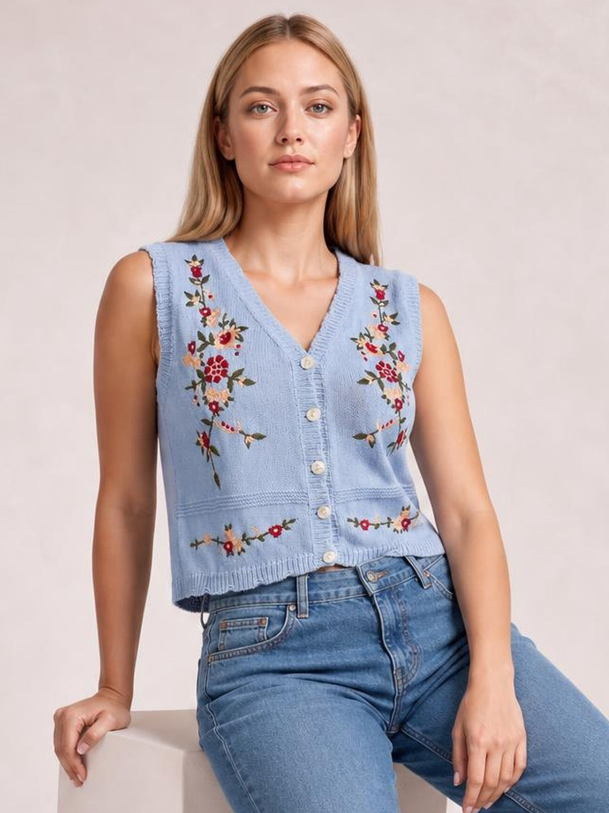 Model seated and facing forward in a light blue embroidered button-up sweater vest with a V-neck, sleeveless cut, floral detailing, and straight silhouette.