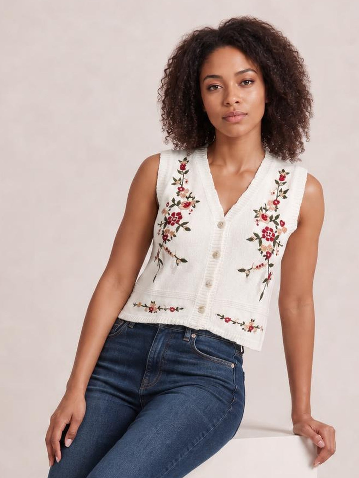 Model seated and leaning back wears a white embroidered button-up sweater vest with a V-neck, floral detailing, sleeveless cut, and cropped silhouette.