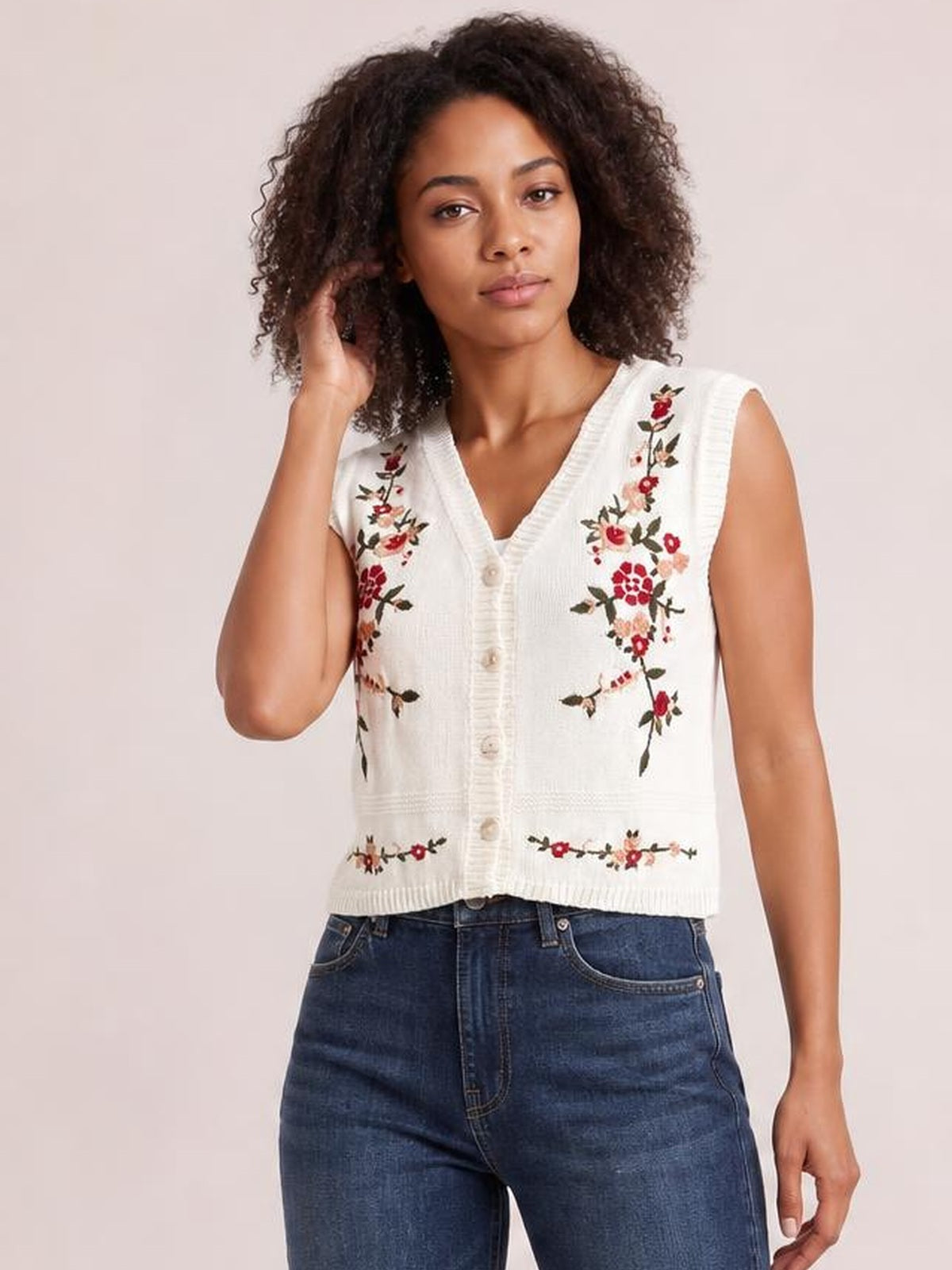 Model wearing a white embroidered button-up sweater vest with a V-neck, floral detailing, ribbed trim, sleeveless cut, and cropped fitted silhouette.