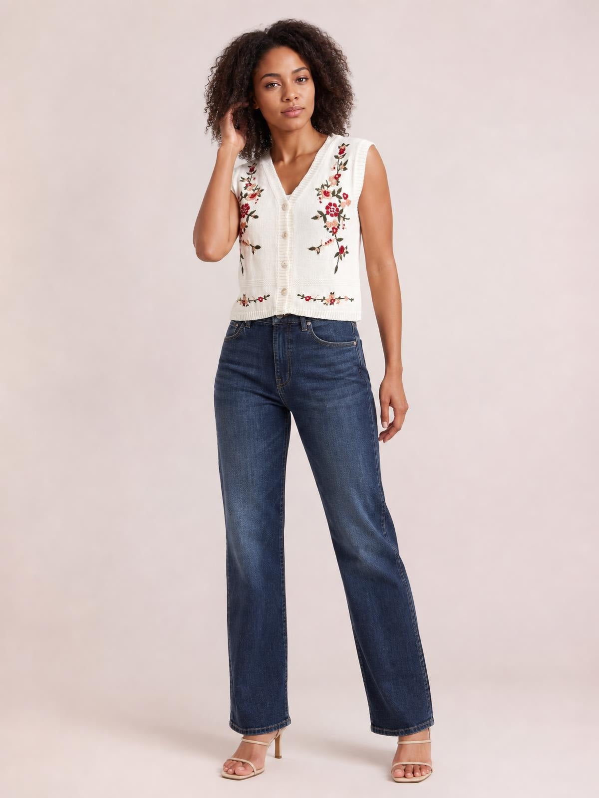Model wearing a cream embroidered button-up sweater vest with a V-neck, sleeveless cut, ribbed trim, and cropped fitted silhouette, posing with one hand in her hair.