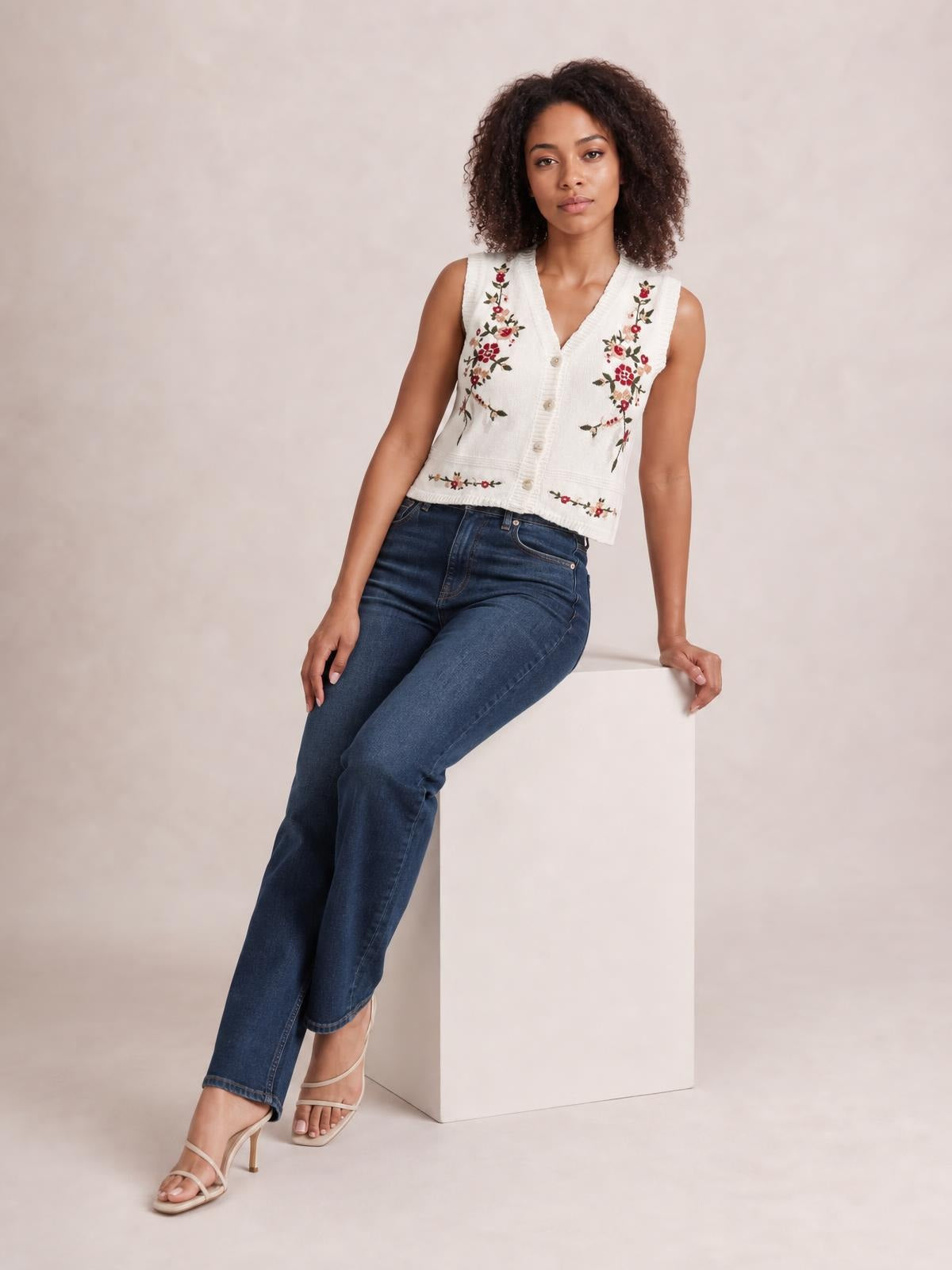 Model seated in a white embroidered button-up sweater vest with a V-neck, sleeveless silhouette, ribbed trim, and front buttons.