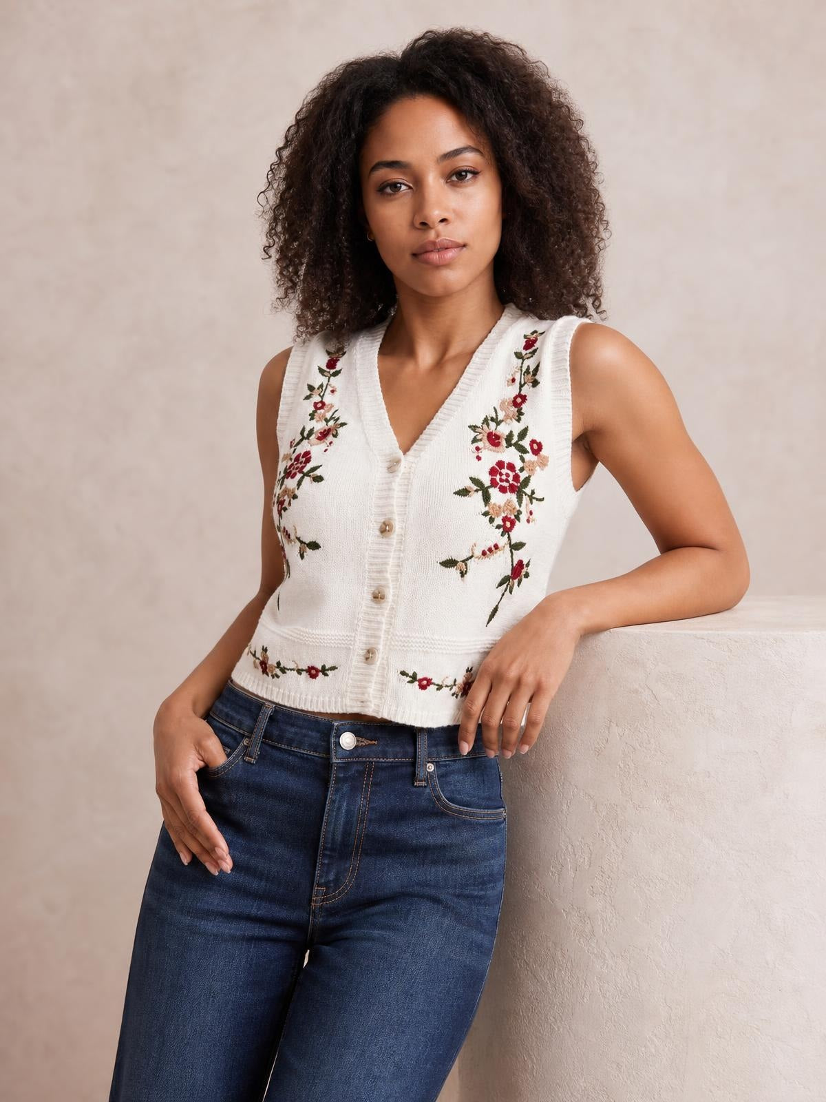 Model wearing a white embroidered button-up sweater vest with floral detailing, ribbed V-neck and hem, fitted sleeveless silhouette, leaning with one arm resting.