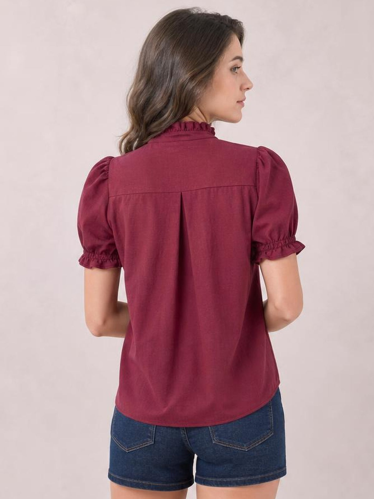 Model shown from the back in a burgundy frill short-sleeve blouse with puffed sleeves, ruffled cuffs, high neckline, back pleat, and curved hem.