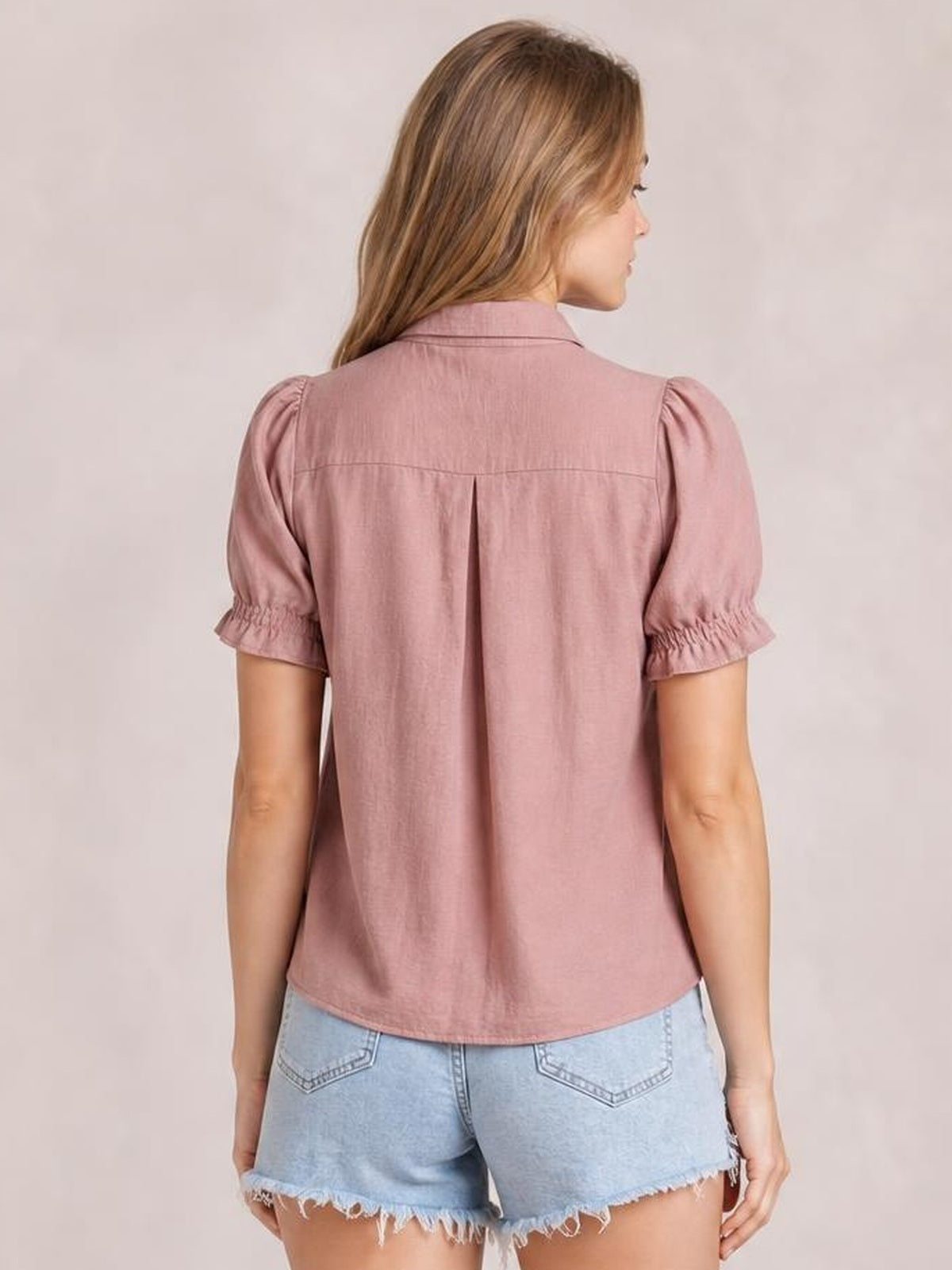 Model shown from behind wearing a dusty pink short-sleeve blouse with a collar, gathered frill cuffs, back yoke, center pleat, and curved hem.