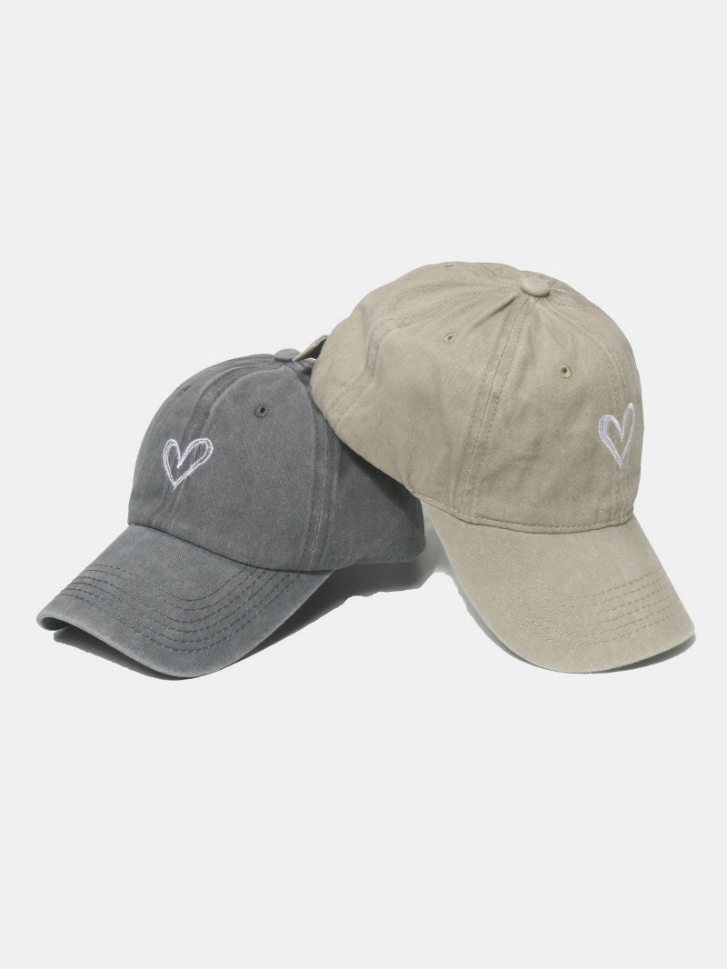 Two washed cotton baseball caps in charcoal gray and beige feature six-panel silhouettes, curved brims, tonal stitching, and embroidered heart motifs.