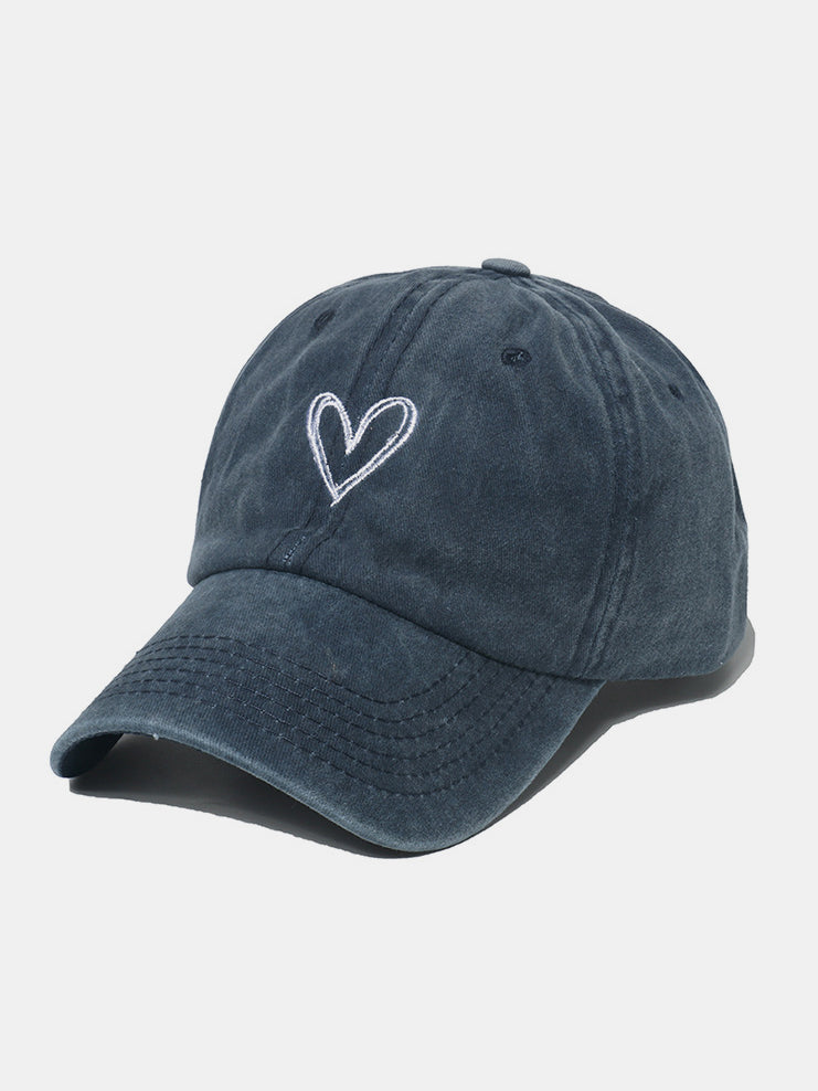 A dark washed cotton baseball cap features a classic six-panel silhouette, curved brim, tonal stitching, and a contrasting embroidered heart on the front.