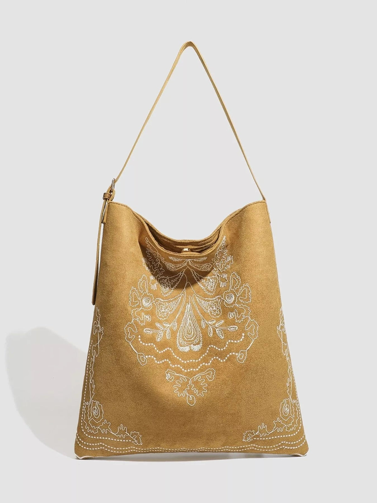 Large tan suede handbag with ornate white embroidery, a slouchy trapezoid silhouette, curved top opening, and a long adjustable shoulder strap.