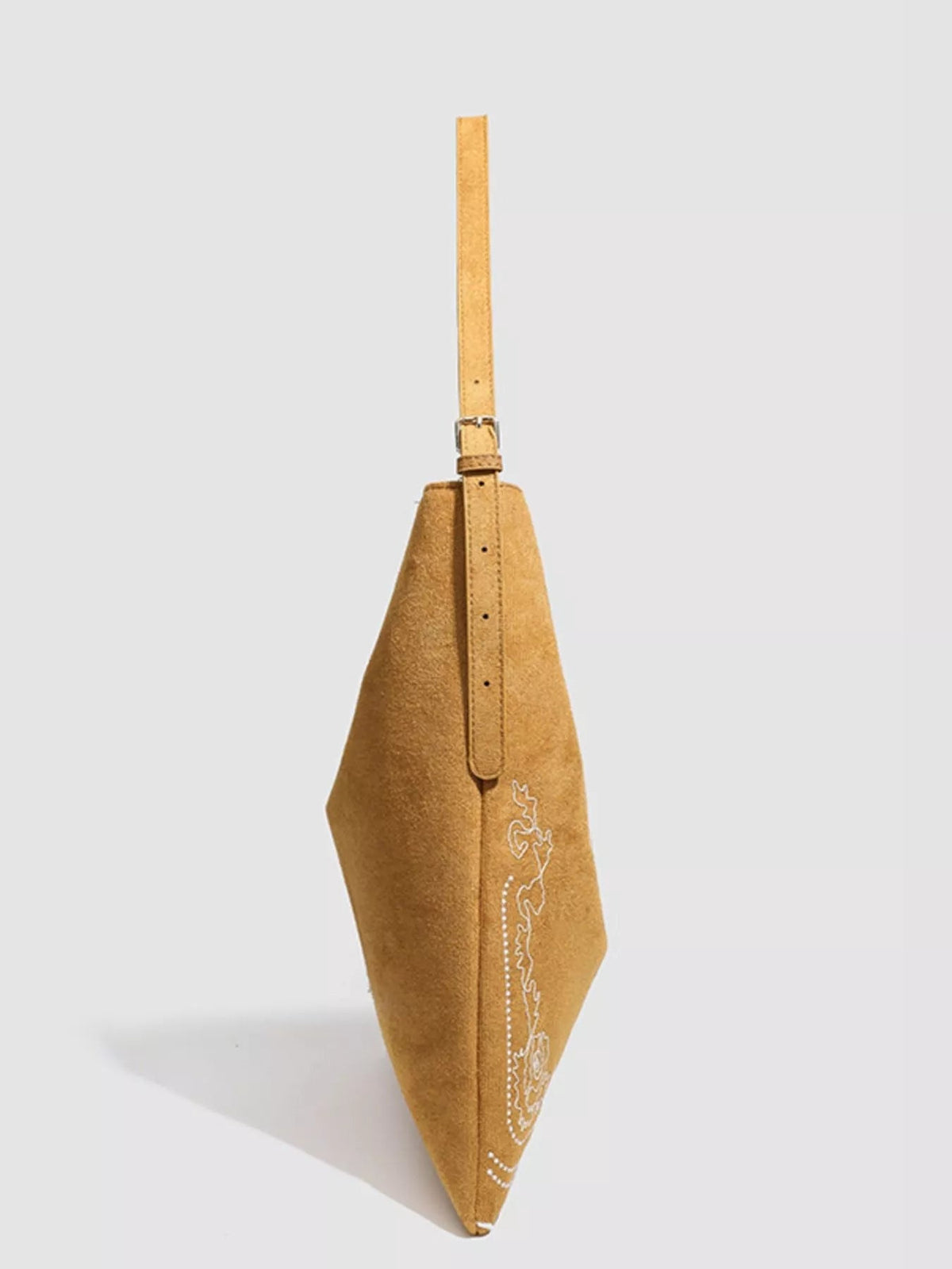 A large tan suede handbag is shown in side profile with a long adjustable strap, tapered silhouette, stitched construction, and white embroidered detailing.