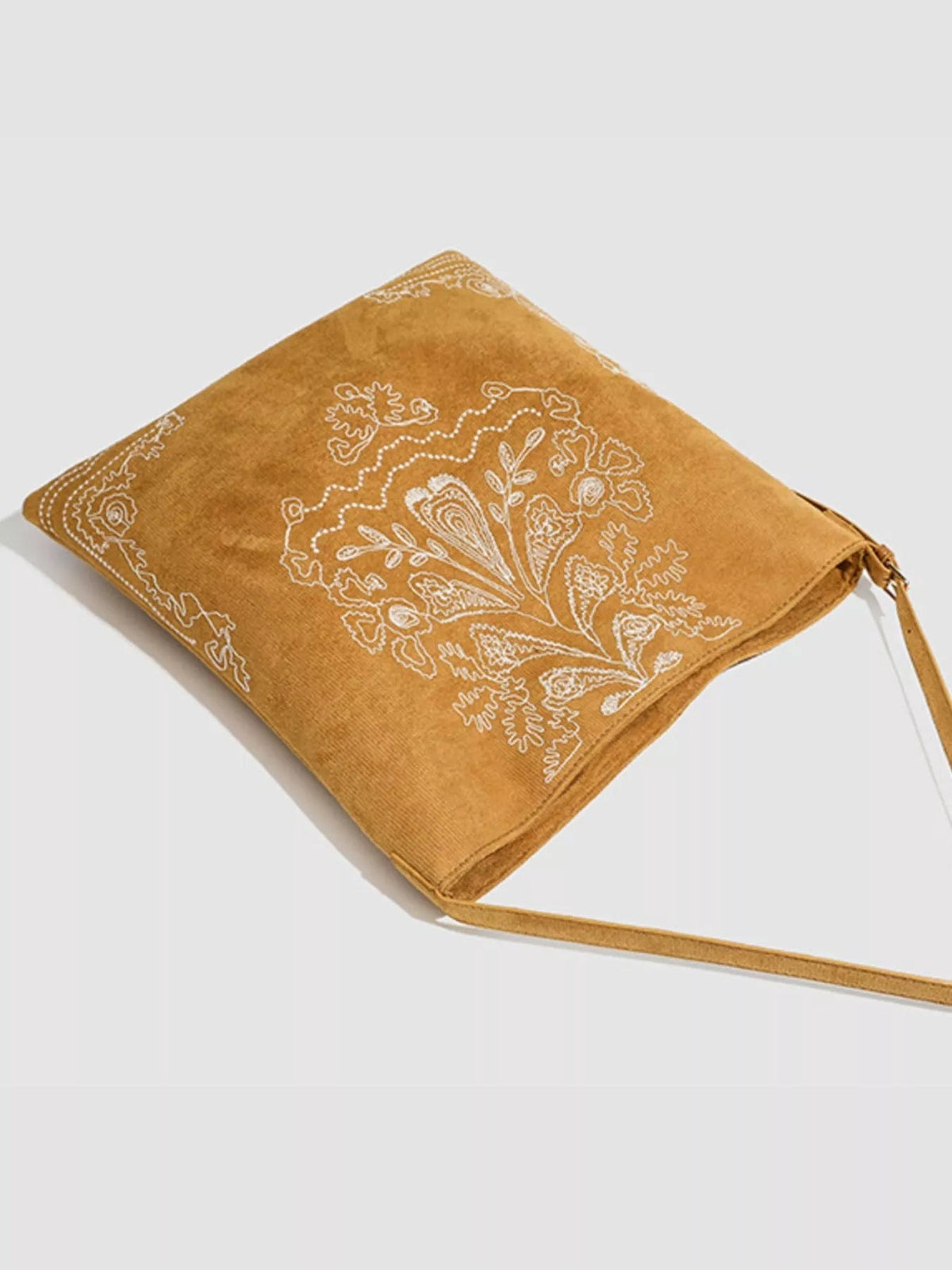A large tan suede handbag features intricate white floral embroidery, a soft rectangular silhouette, and a long slender strap.