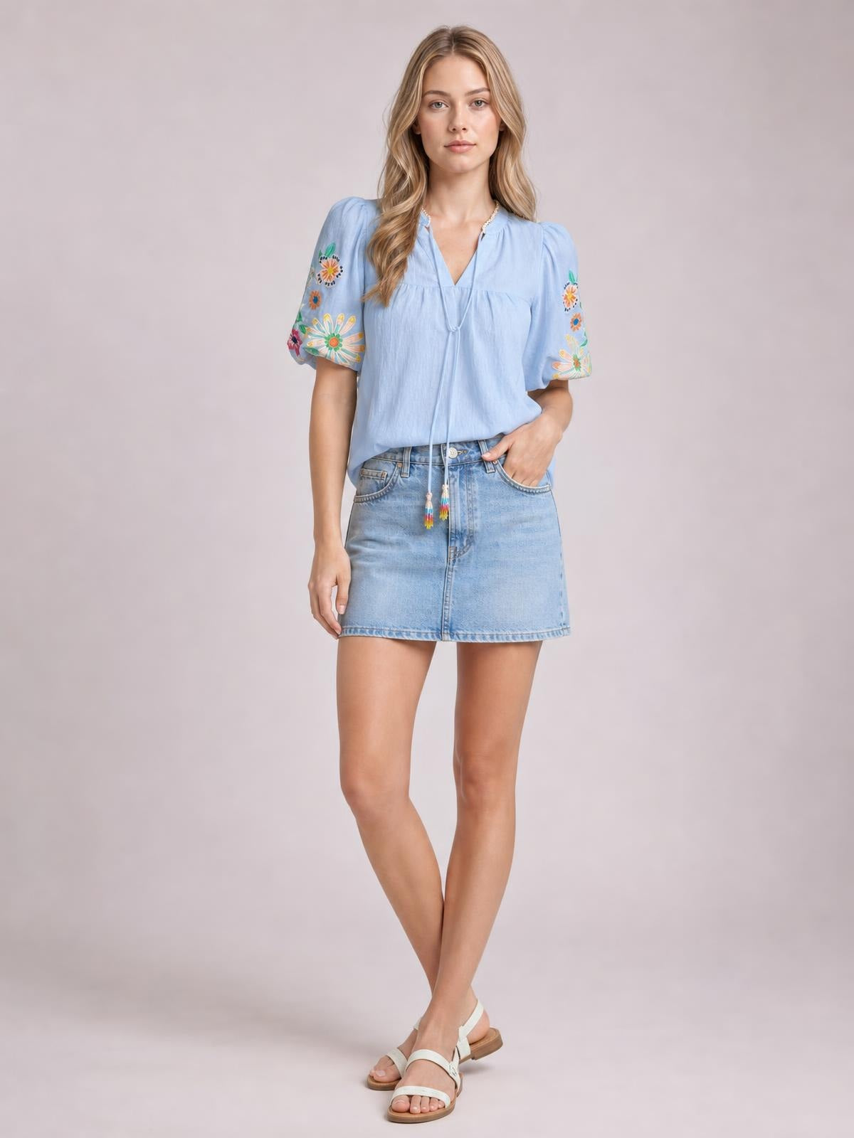 A model stands with one hand in her pocket wearing a light blue embroidered tie neck blouse with short puff sleeves and a relaxed silhouette.