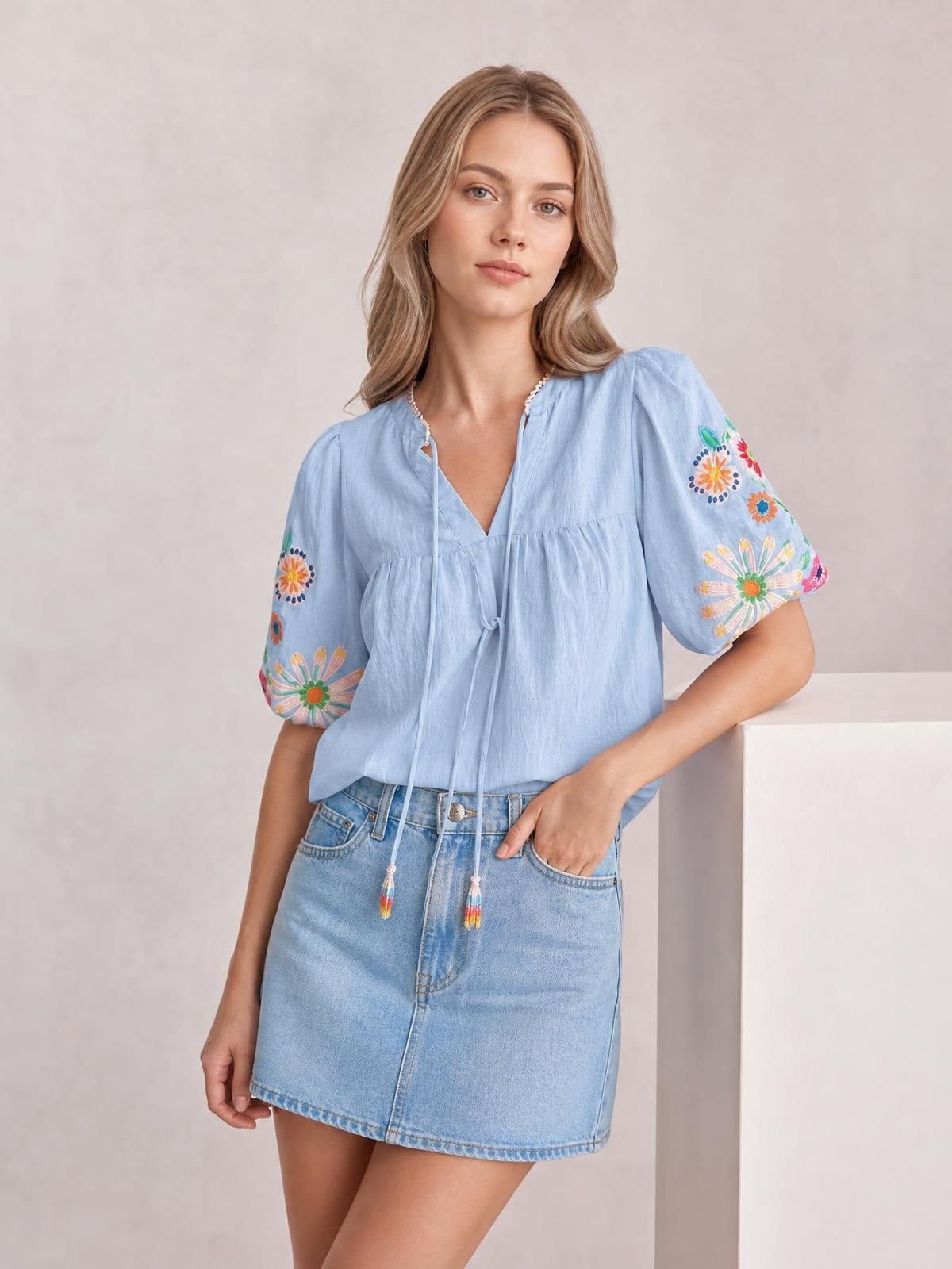 Model poses with one hand in a pocket wearing a light blue embroidered tie-neck blouse with short puff sleeves and a relaxed, gathered silhouette.