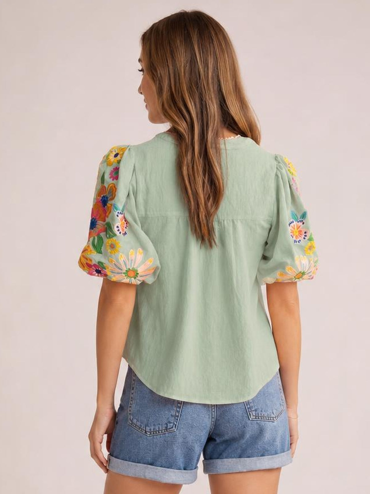 Model shown from behind wearing a mint green embroidered blouse with short puff sleeves, colorful floral stitching, a relaxed silhouette, and curved hem.