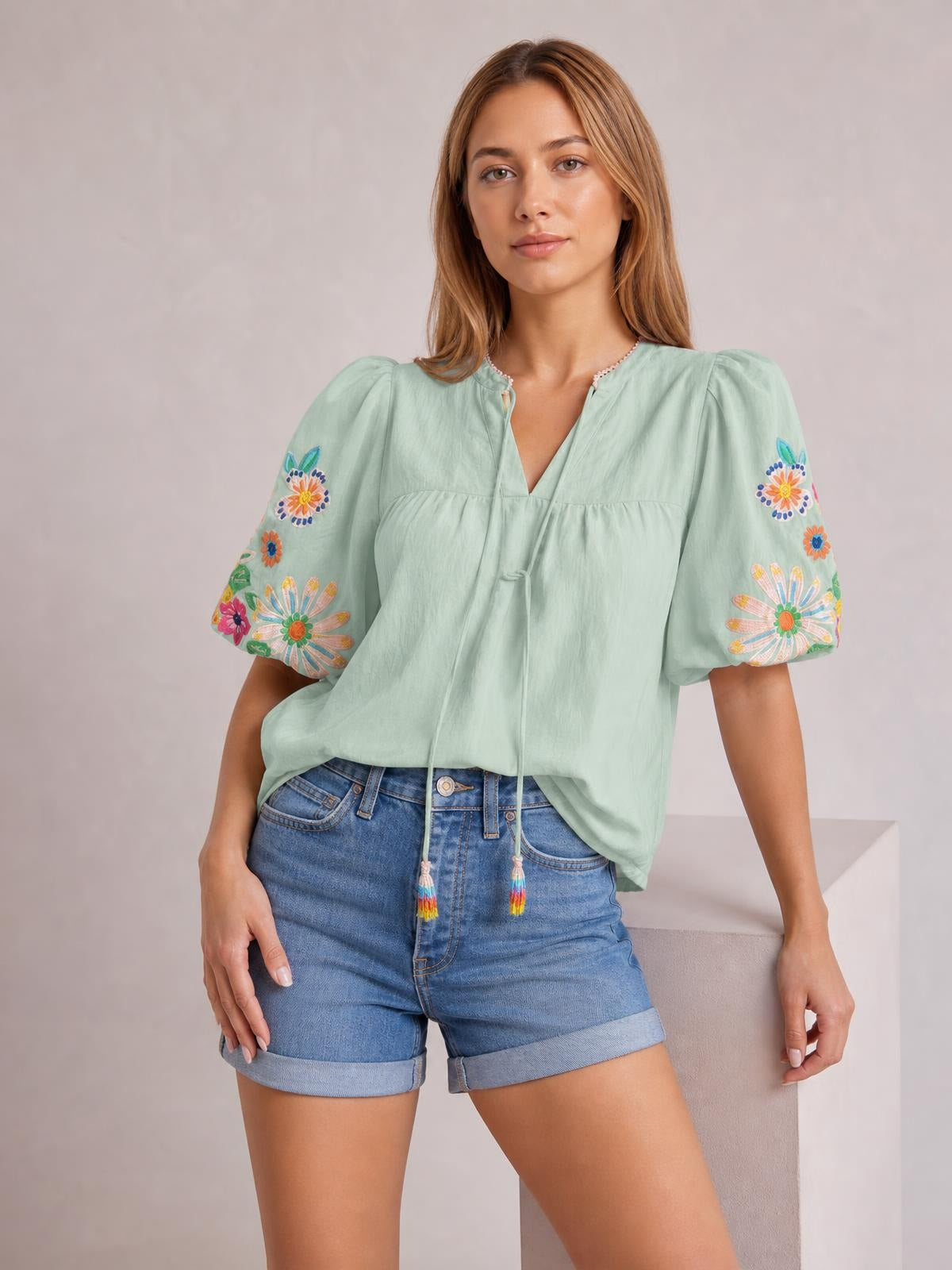 A model stands in a mint green embroidered tie-neck blouse with short puff sleeves, floral sleeve detailing, a V-neckline, and a relaxed gathered silhouette.