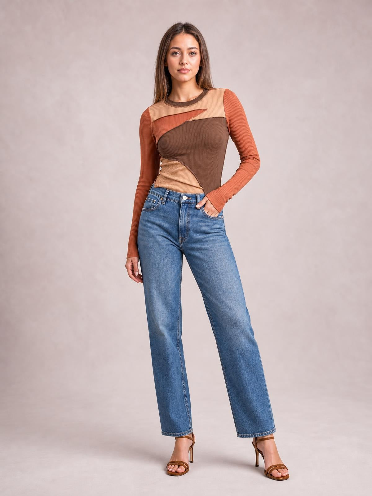 Model poses with one hand in her jeans pocket wearing a fitted contrast round-neck long-sleeve T-shirt in rust, tan, and brown with exposed diagonal seams.