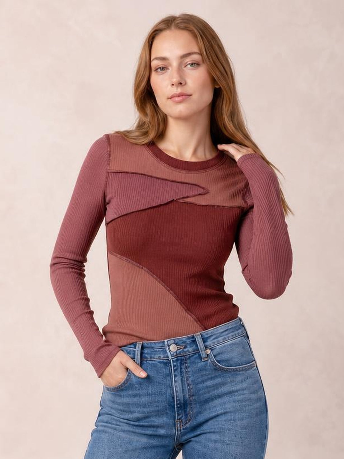A model poses in a fitted mauve and burgundy contrast long-sleeve T-shirt with a round neck, exposed seams, ribbed texture, and curved panel construction.