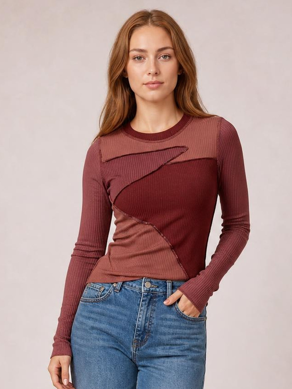 A model poses in a fitted burgundy contrast round neck long sleeve T-shirt with exposed diagonal seams and a ribbed texture, one hand in her jeans pocket.