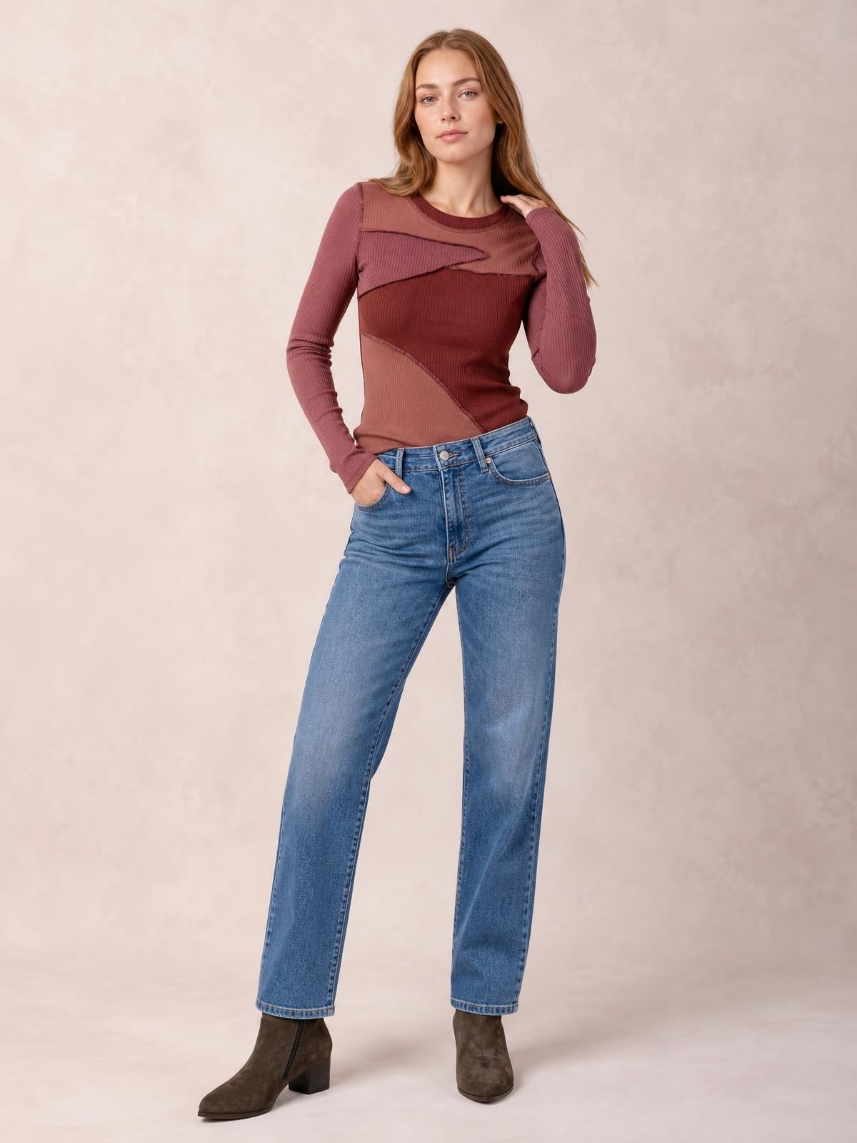 Model wears a burgundy long-sleeve round-neck T-shirt with contrasting exposed seams and a fitted silhouette, posing with one hand in her jeans pocket.